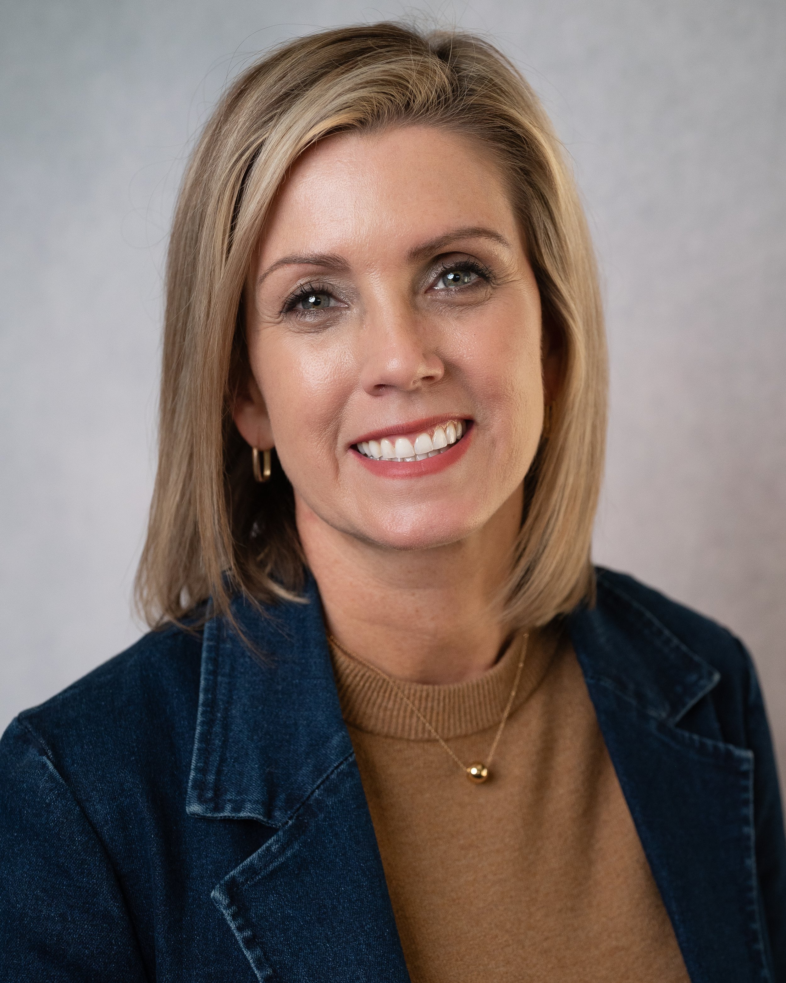 A woman with shoulder-length blonde hair, wearing a green knitted top with small perforations. She is smiling, has gold hoop earrings, and a gold necklace with a pendant. The background is plain and neutral.