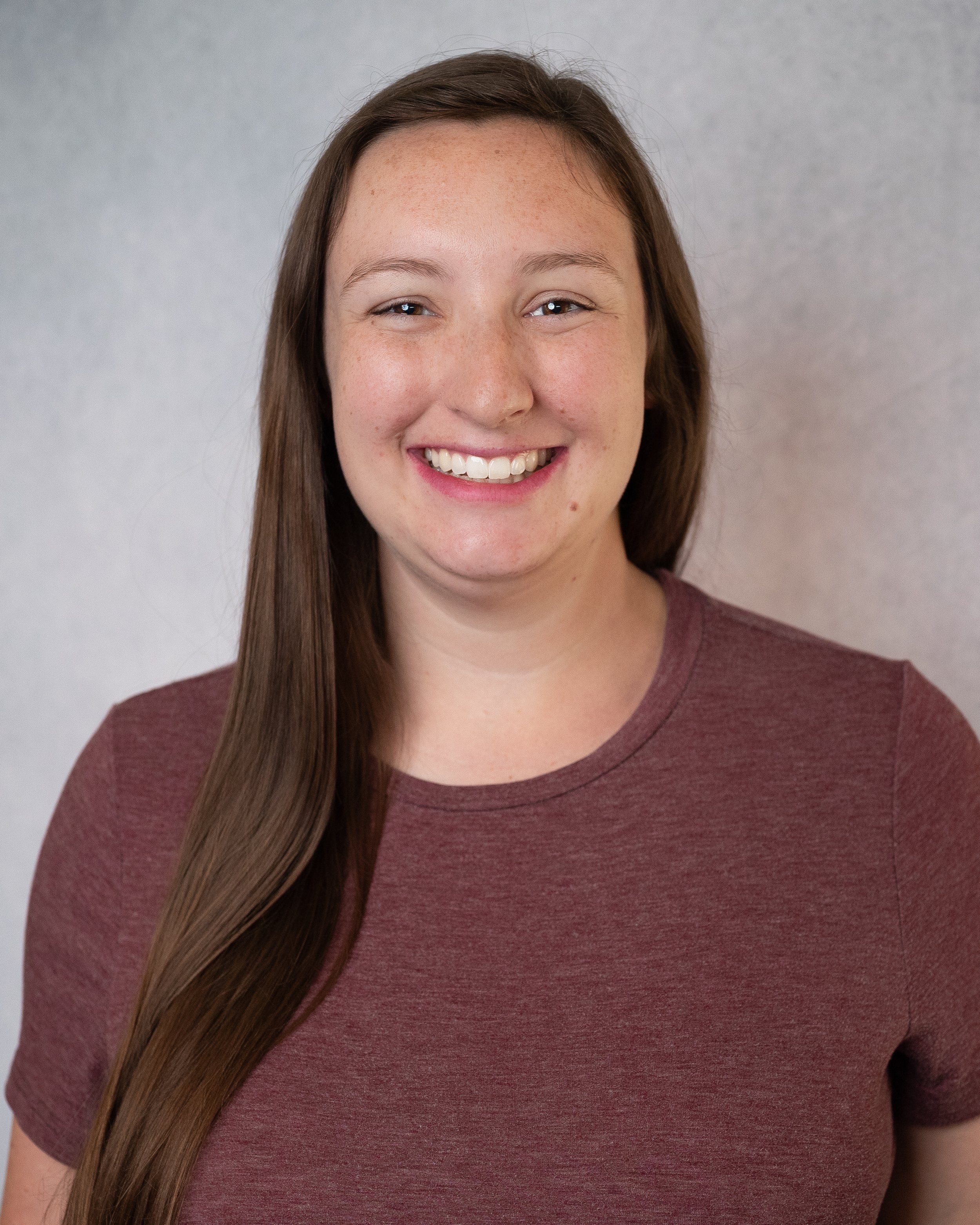 A smiling woman with long brown hair and freckles wearing a dark gray top, against a green background.