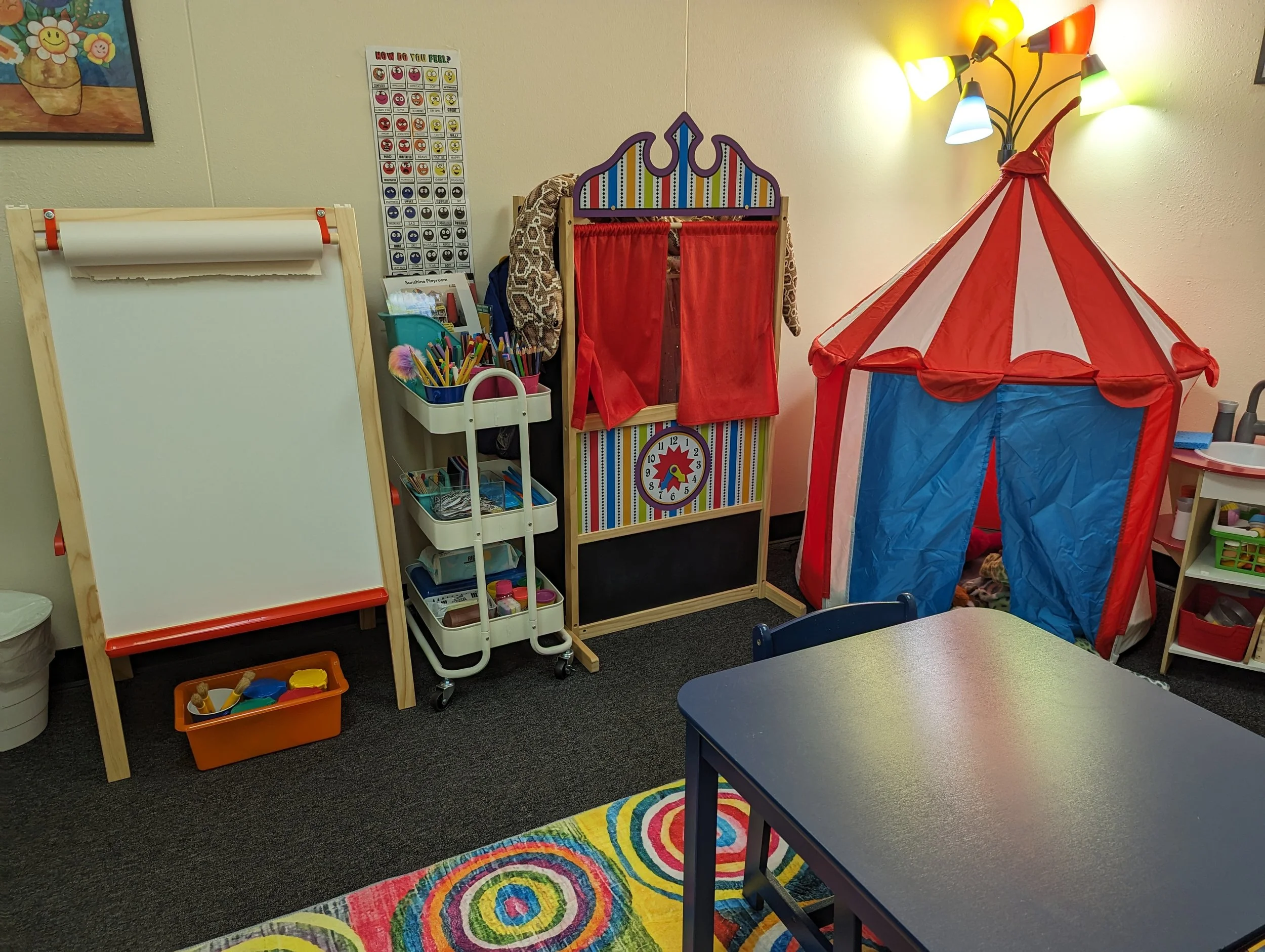 Children's play area with a red and blue play tent, colorful carpet, whiteboard on stand, art supplies, toys, and decorative wall posters.