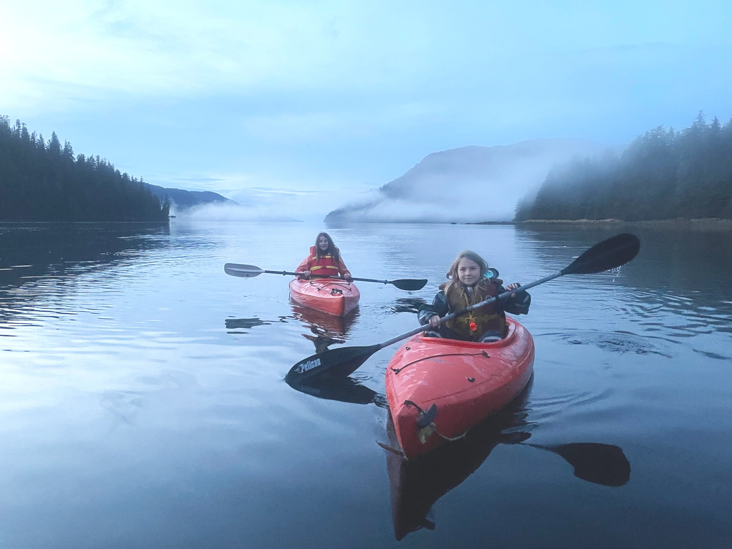 Two young girls kayaking in red kayaks at dusk.  Low clouds and mist covering hills and trees in the background.