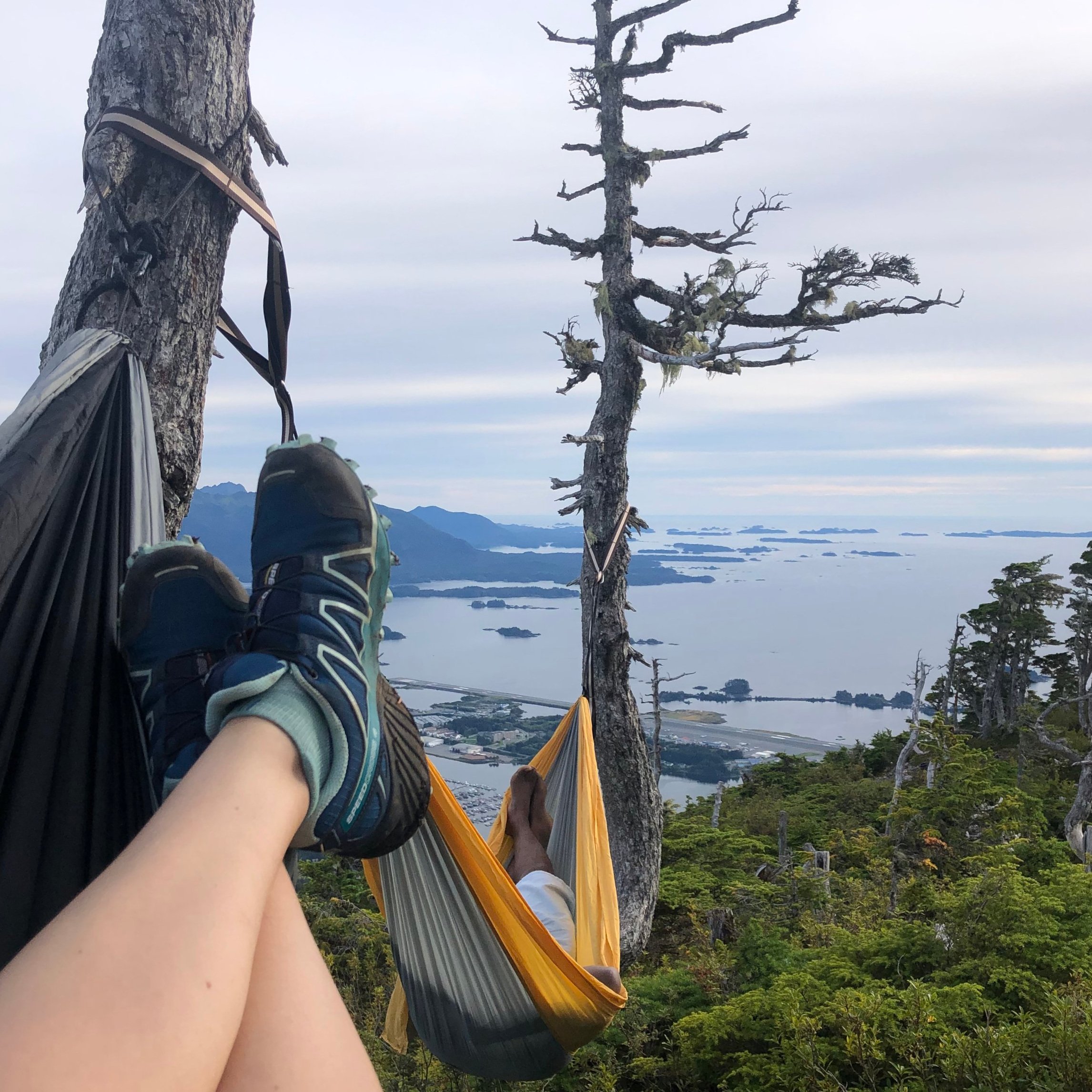 Photo taken by person laying in hammock set up in the alpine, legs and shoes showing with standing dead tree and ocean and islands beyond.
