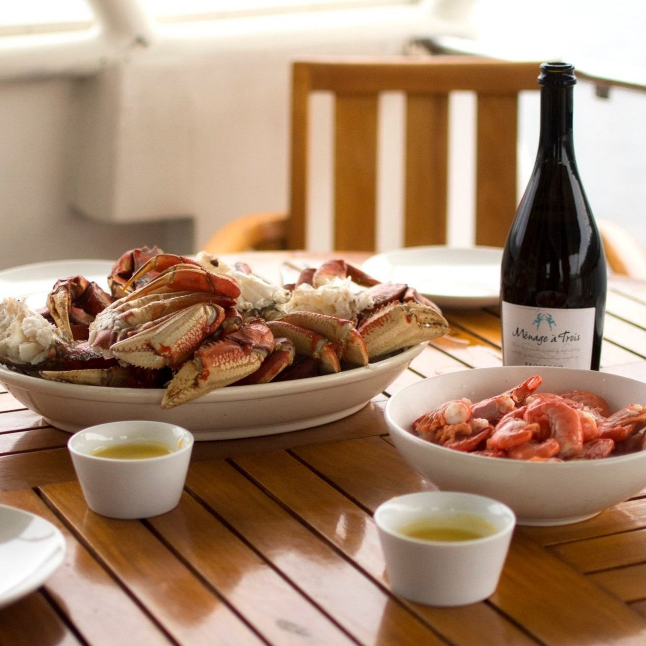 A table setting with platters full of Dungeness crab and prawns, with a bottle of wine and small dishes of melted butter.