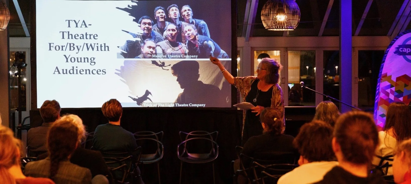 A woman speaking to a conference audience, with a screen showing images of theatre for young audiences.