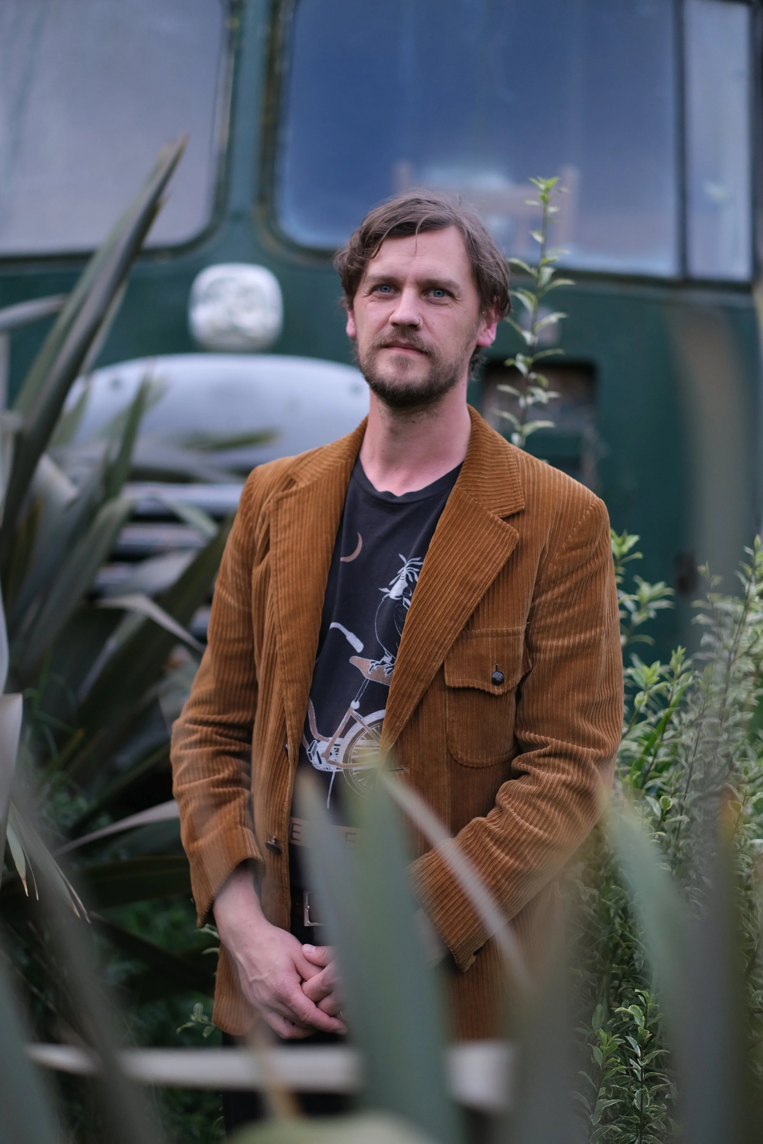 A man with brown hair and a beard, wearing a brown corduroy blazer and a black graphic t-shirt, stands outdoors among green plants with a dark green vehicle in the background.