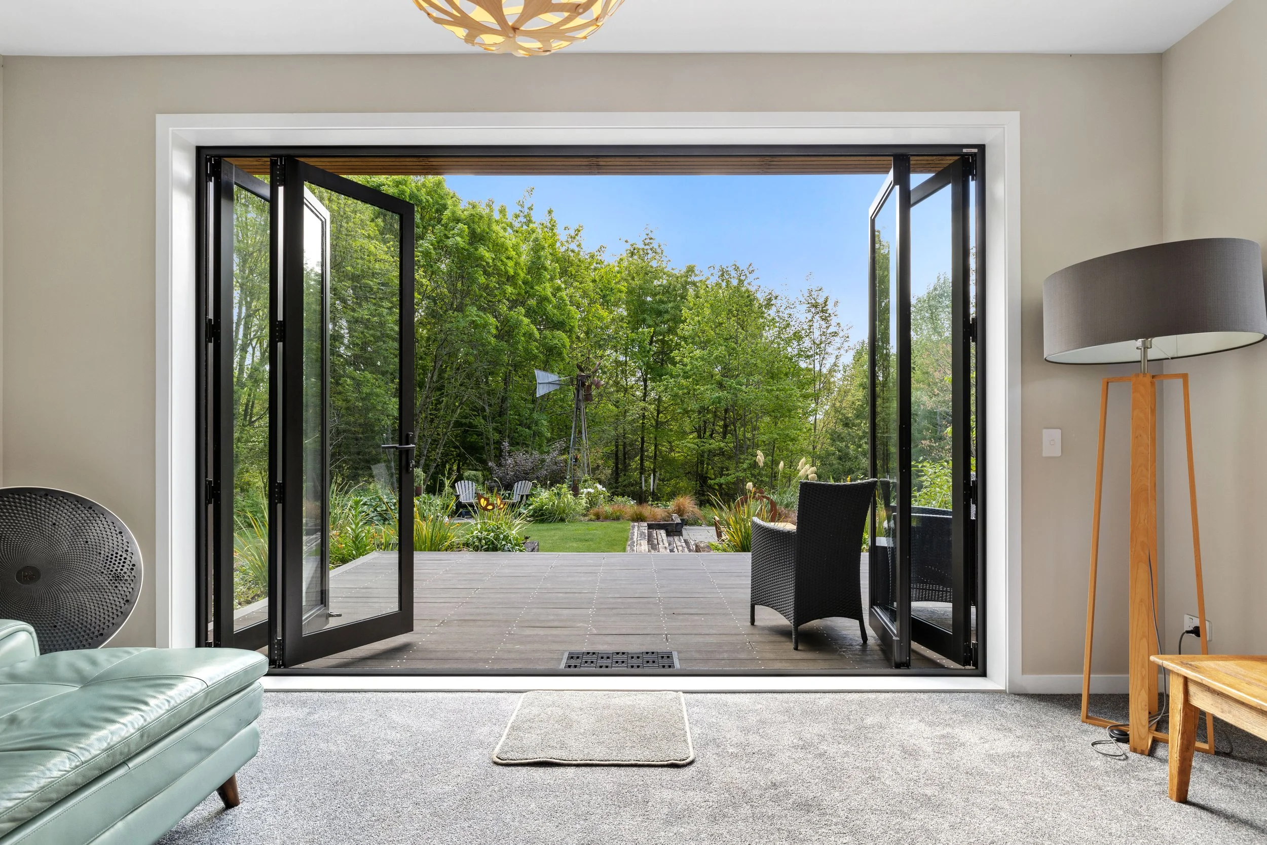 Open glass doors leading to a backyard garden with trees, chairs, and a windmill, viewed from inside a room with beige walls, gray carpet, a floor lamp, and a small wooden table.