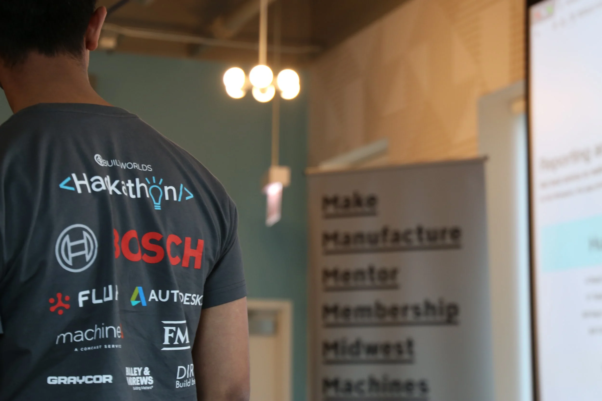 Person wearing a gray t-shirt with hackathon and sponsor logos, including Bosch and Autodesk, in a conference setting.