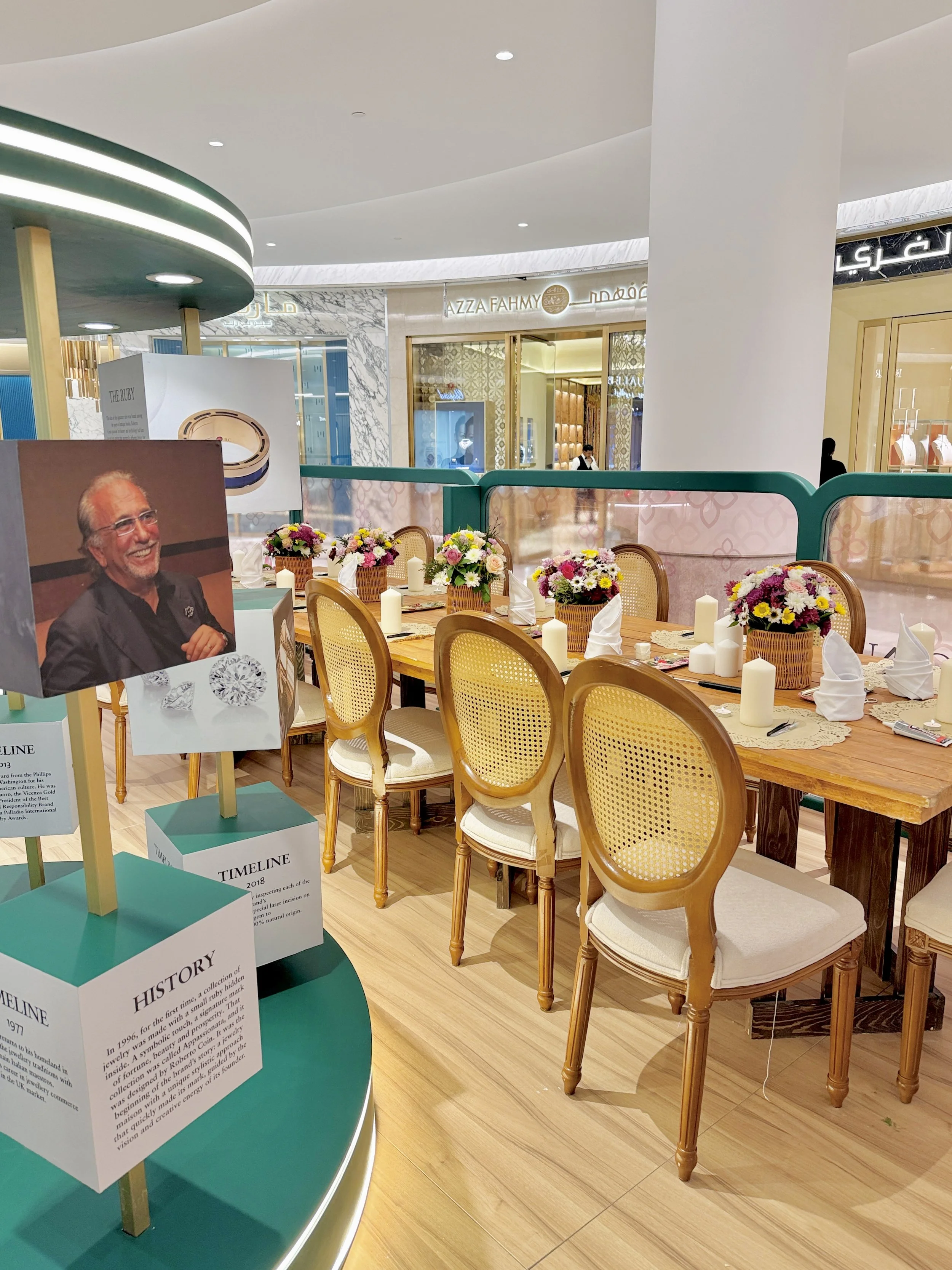 A display table with informational cards and a backdrop of a dining area inside a shopping mall. The dining area features wooden chairs with cane backs and a long wooden table decorated with flower arrangements and white candles. In the background, t
