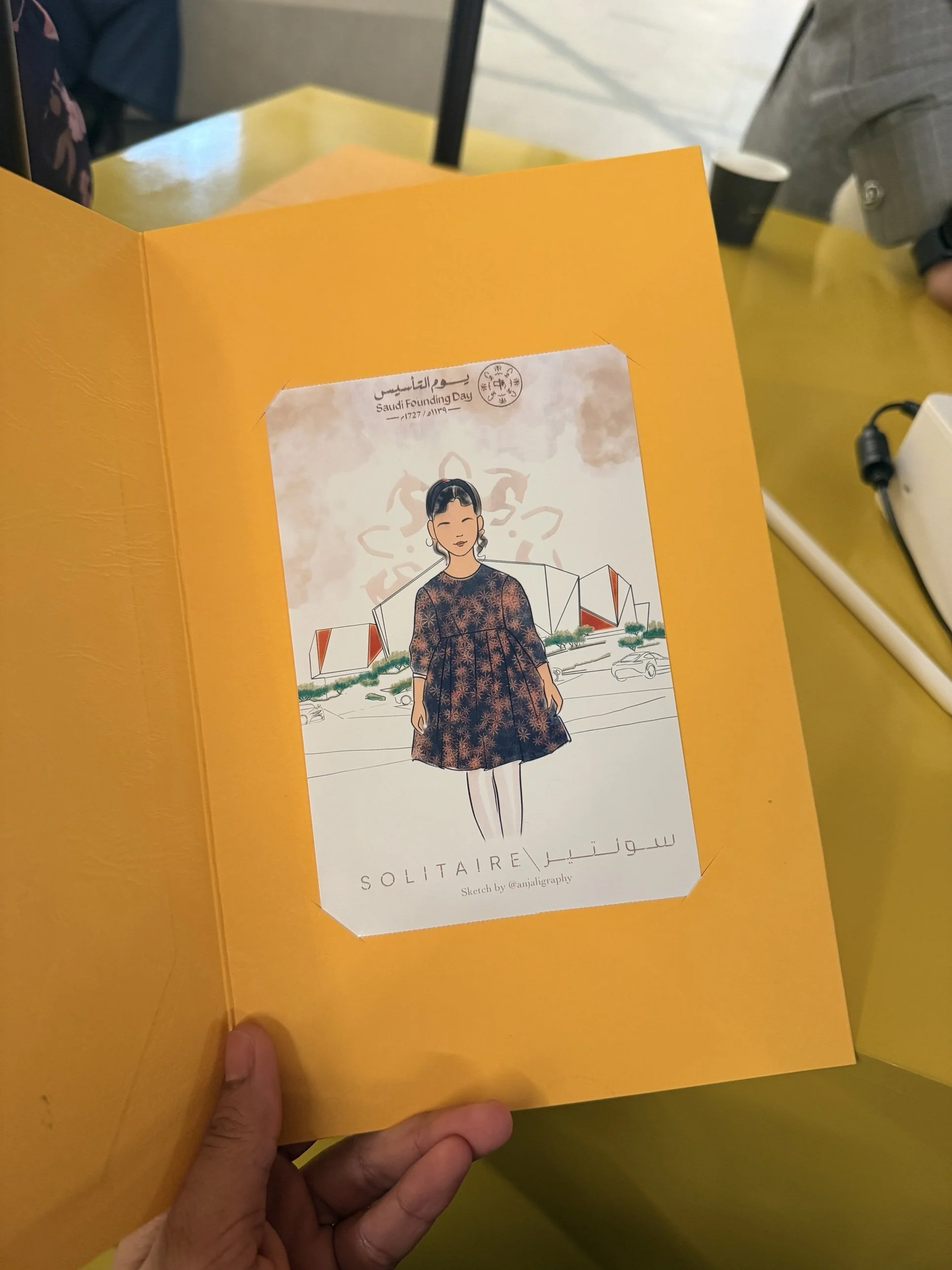 A hand holding an orange folder with a drawing of a woman in a patterned dress on a white paper sheet inside the folder, with text 'SAUDIARABIA SOLITAIRE' and 'Sketch by @anjalligraphy' at the bottom.