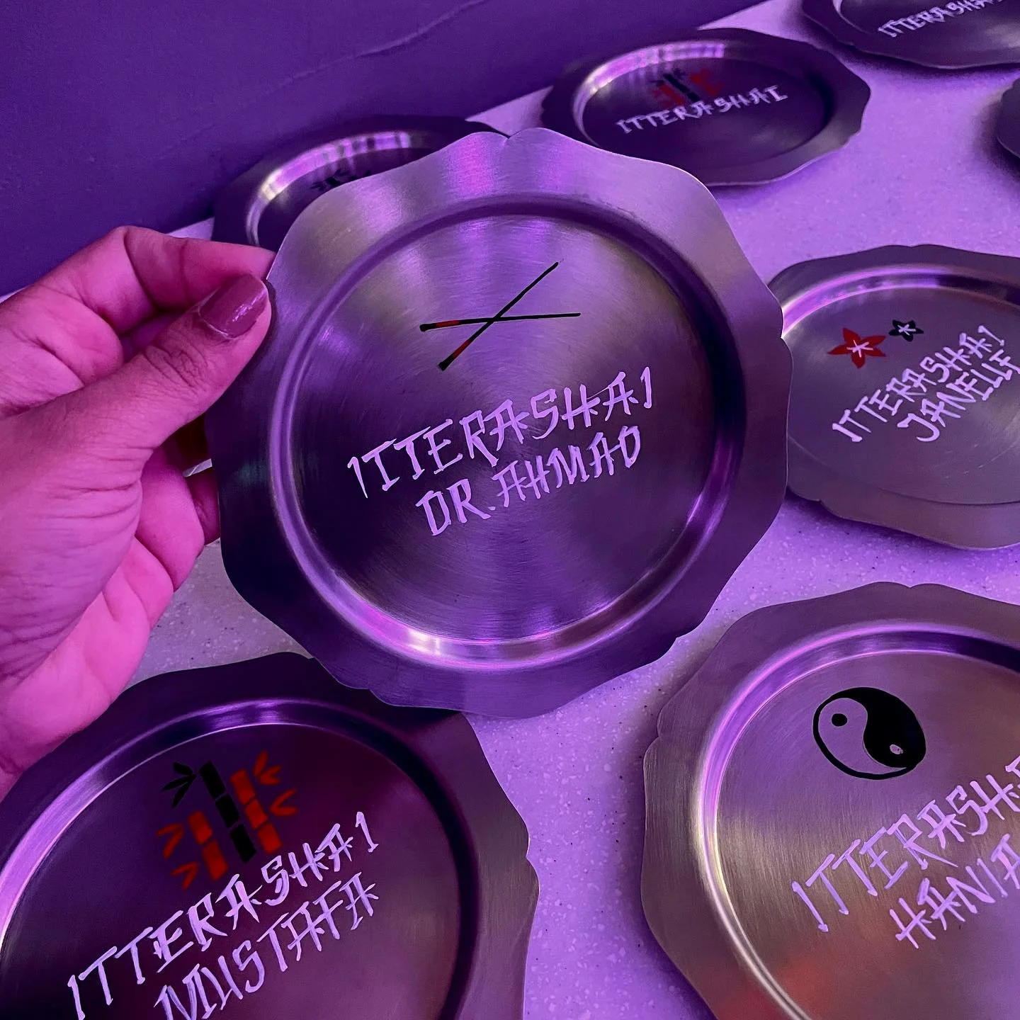 Decorative metal trays with the names "Iterationsha Janelle" and various symbols, including a clock and yin-yang, in a setting with purple lighting.