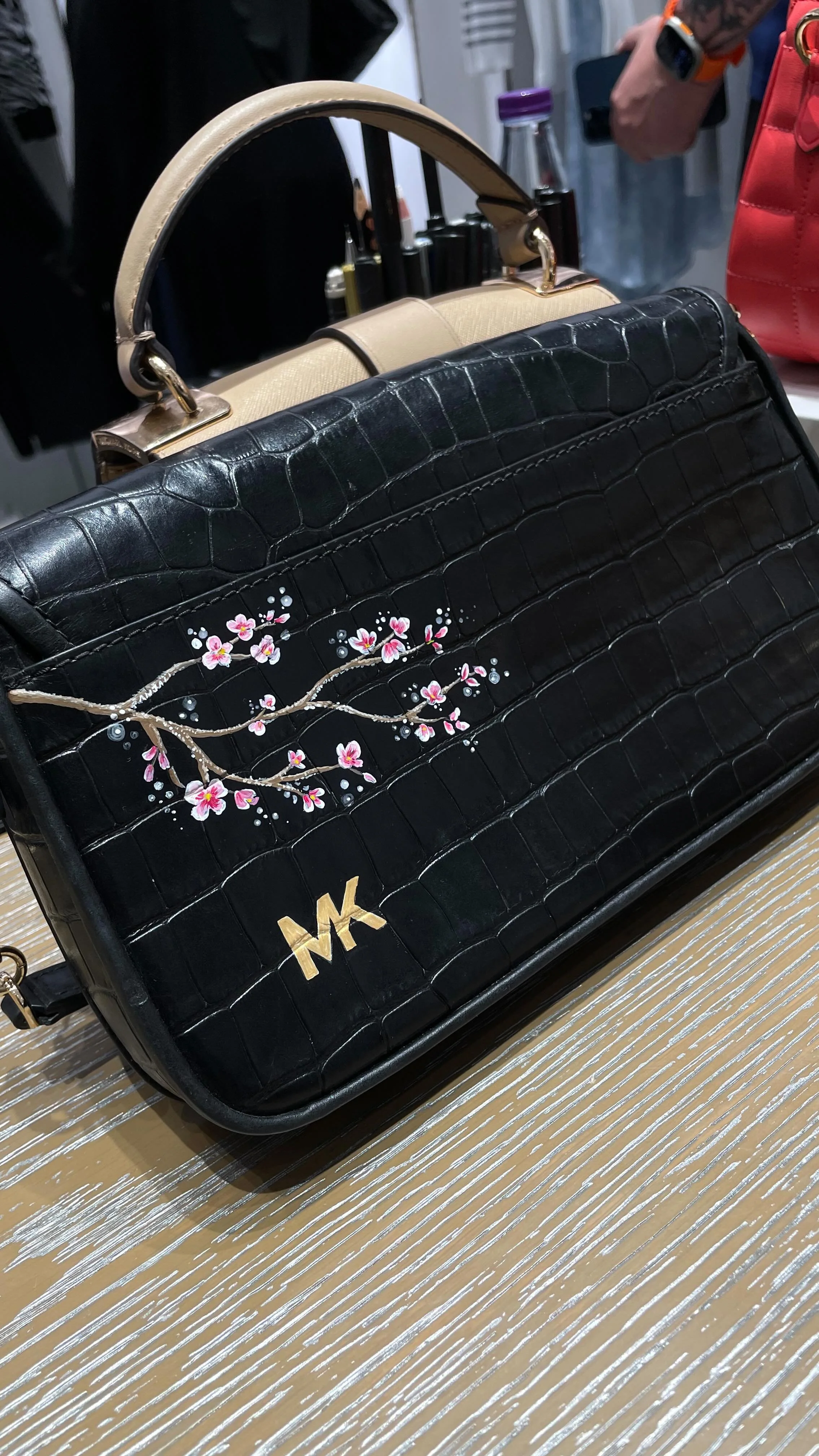 A black designer handbag with a floral branch design, gold initials, and a beige handle, placed on a light-colored wooden surface.