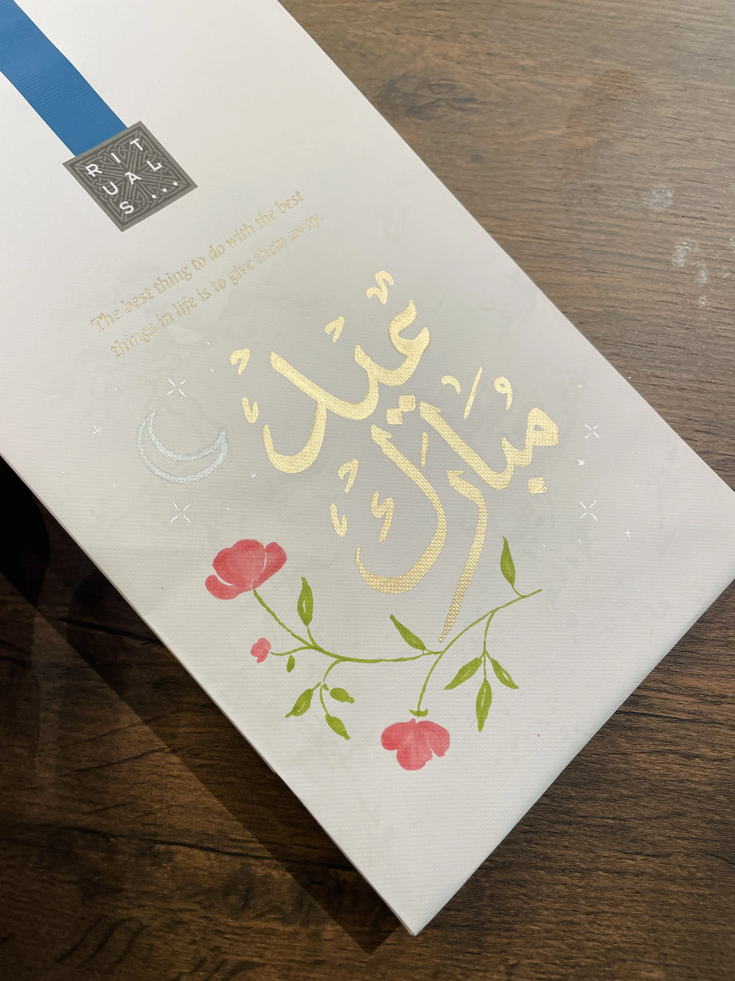 A white piece of paper with gold Arabic calligraphy and floral design, also features a small quote in English reading 'The best thing to do with the best things in life is to give them away'.