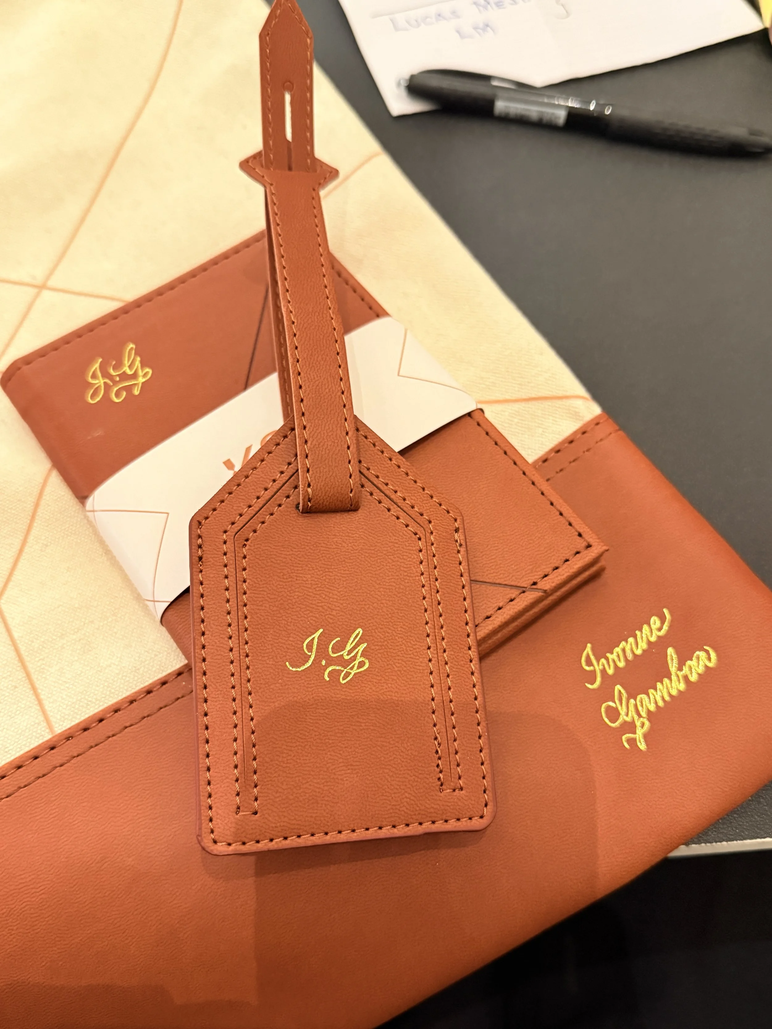 Close-up of a tan leather gift bag with a matching luggage tag, and a gold-embossed logo that reads "J. Crew". There are papers and a pen on a black surface in the background.