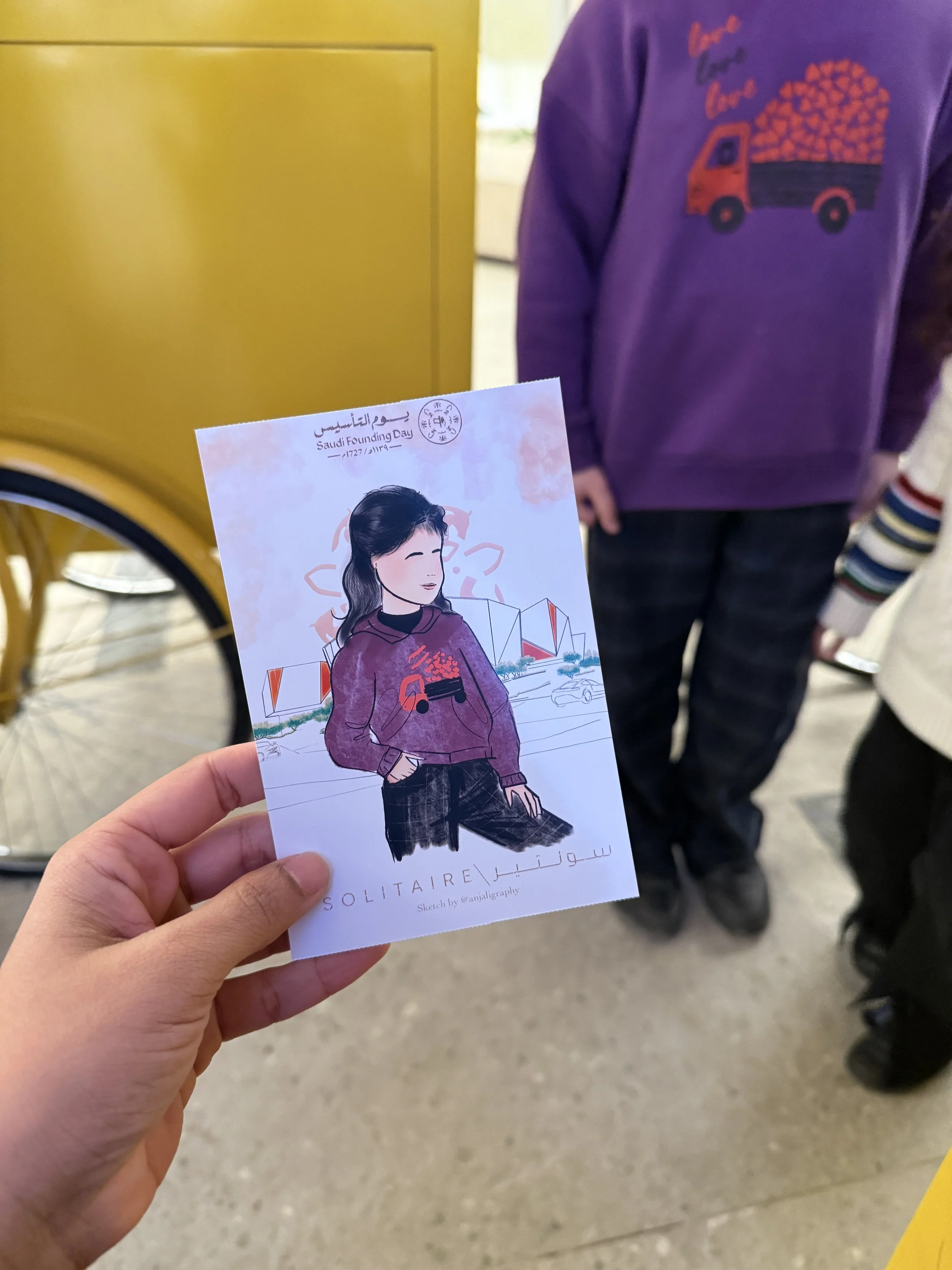 A person holding a color illustration of a woman in a purple jacket, with a background showing a yellow cart and other people.