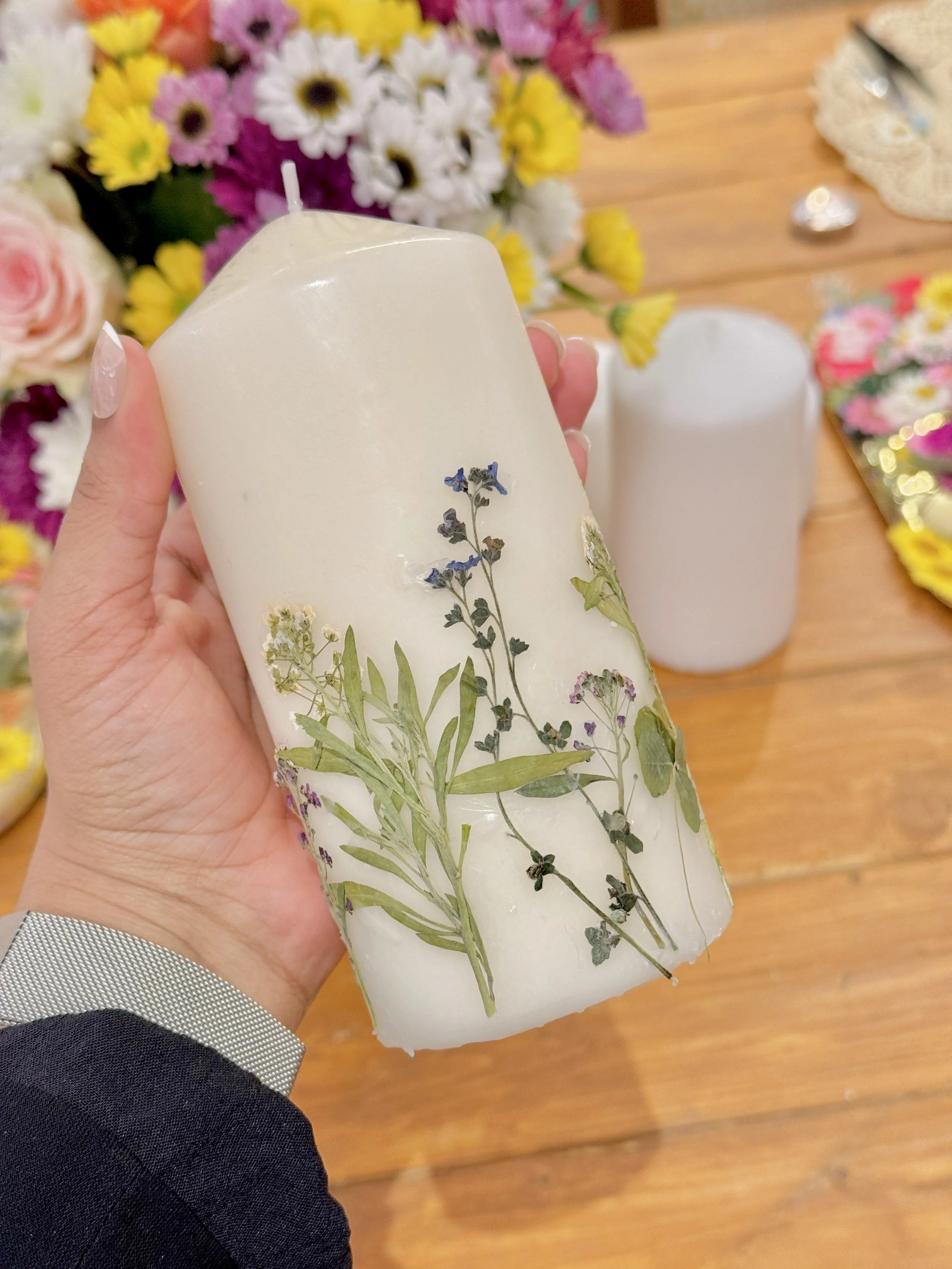 A hand holding a large white candle decorated with pressed lavender and other small flowers, with a background of colorful flowers and a table.