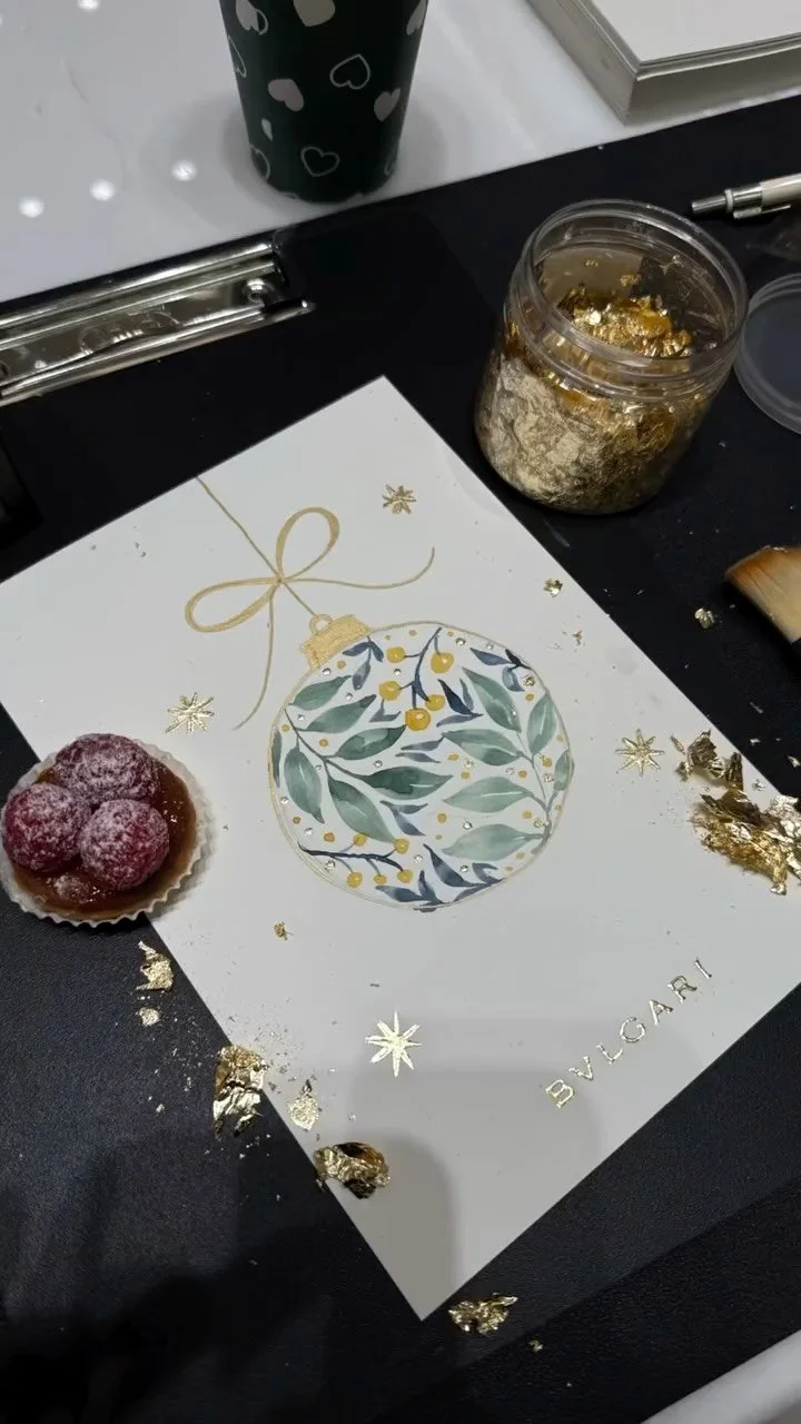 A holiday-themed card with gold accents, a jar of gold flakes, a black cup with white hearts, a pen, a small tart with raspberries and powdered sugar, and gold foil scattered around.