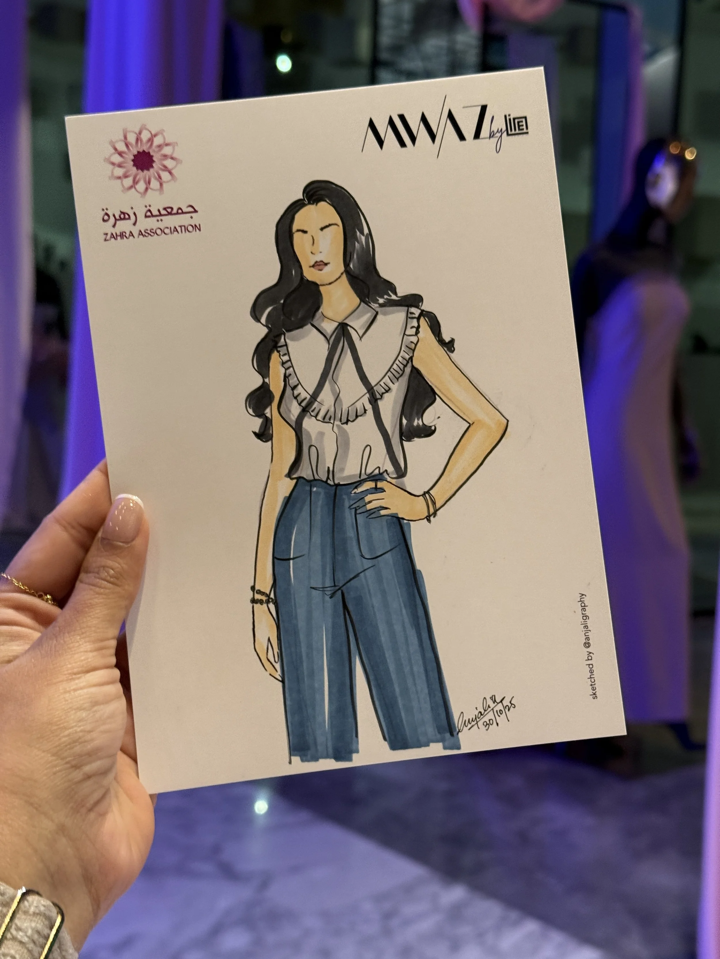 A hand holds a sketch of a woman wearing high-waisted pants and a sleeveless blouse, with long dark hair. The drawing is on a piece of paper with logos and text in Arabic and English, including 'Zahra Association' and 'MWAZ by Liel.' The background s