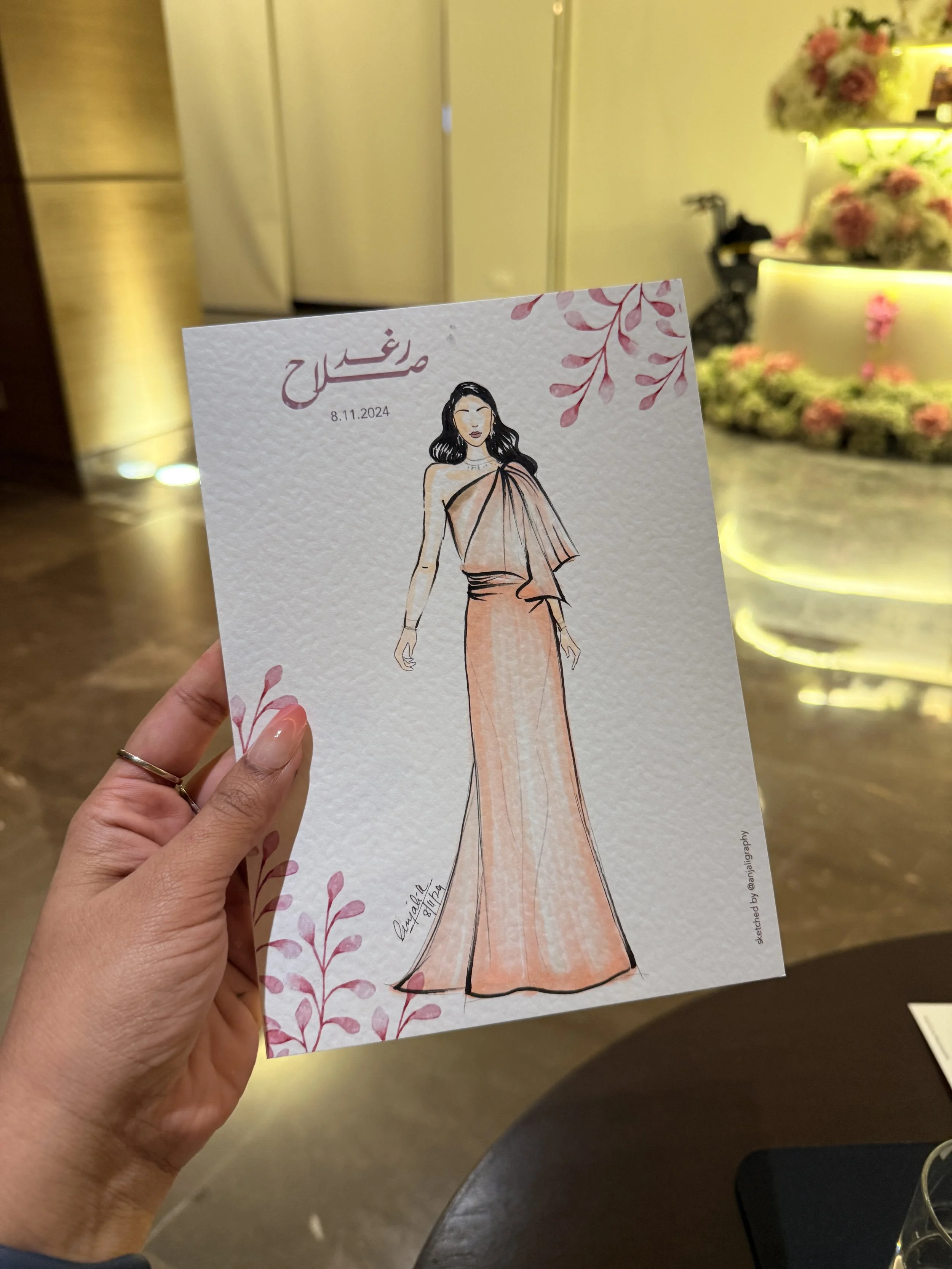 A hand holding a wedding invitation card with an illustration of a woman in a peach-colored dress, and floral decorations on the card. The background shows a floral arrangement and a soft glowing light.