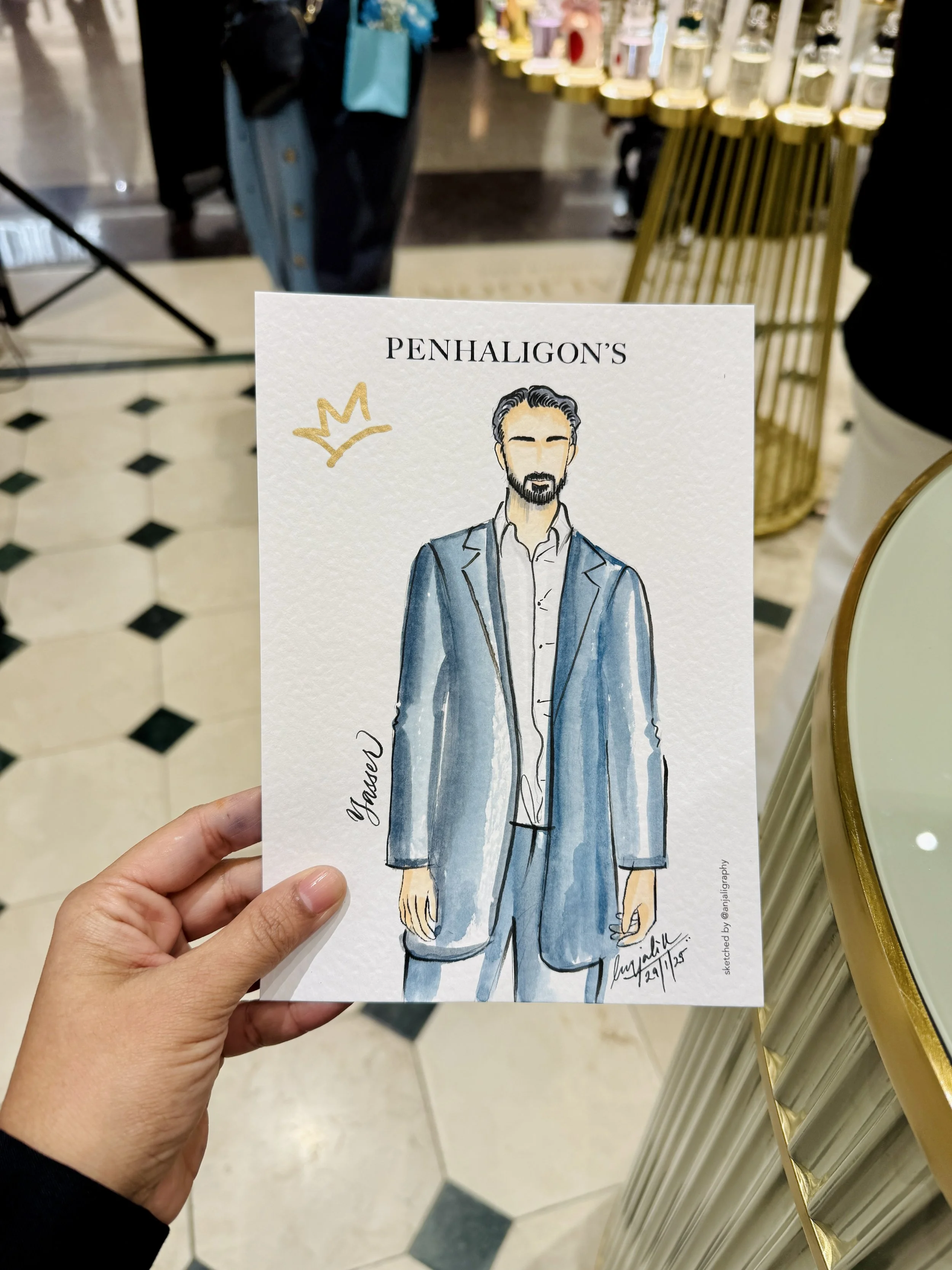 A hand holding a sketch of a man in a suit with a white shirt and blue blazer. The sketch is signed with the date 29/7/25 and the artist's Instagram handle. The background shows a store with perfume bottles and a tiled floor.