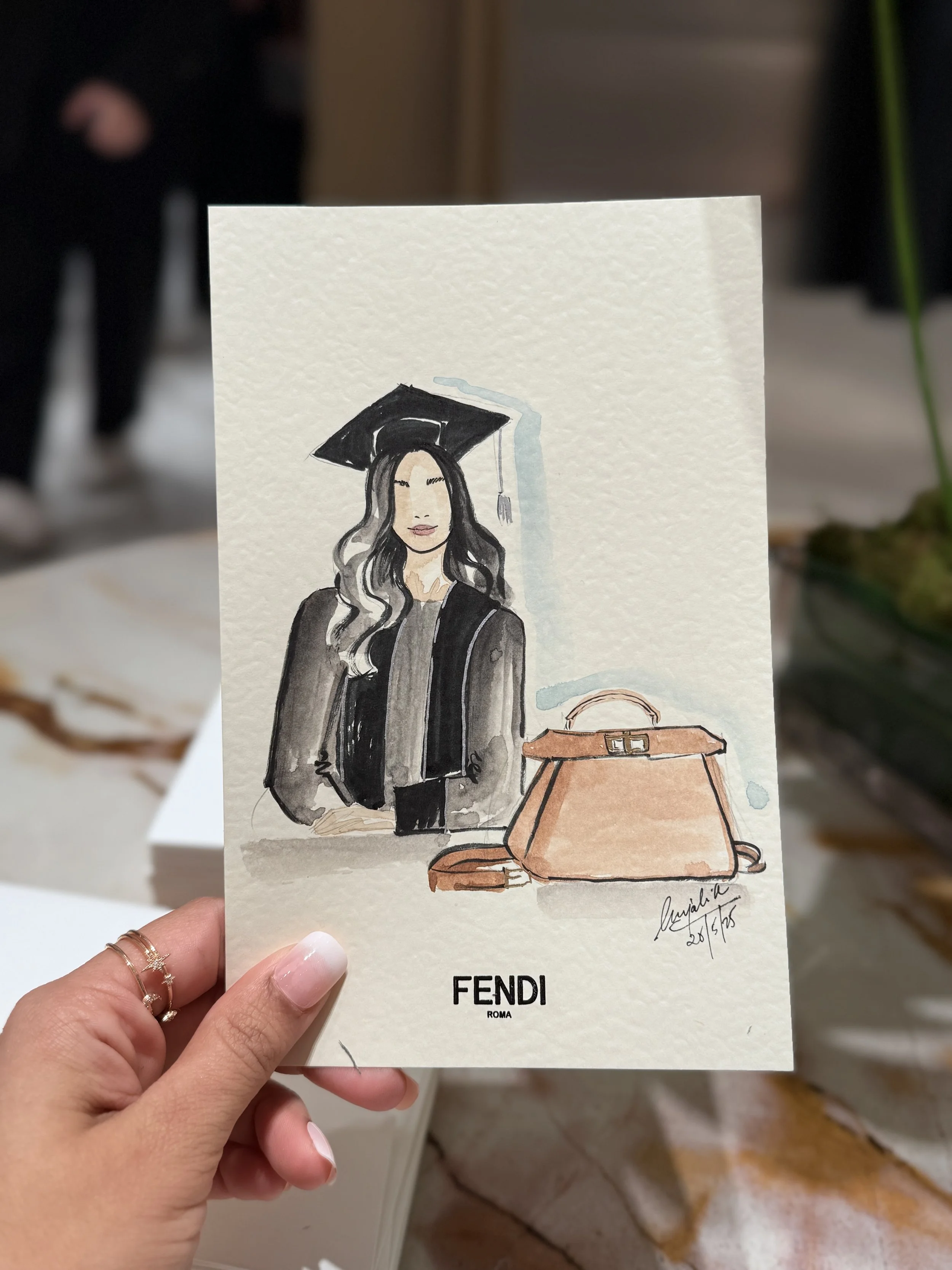 Colorful watercolor illustration of a woman with long hair wearing a graduation cap and black gown, with a brown handbag placed on a table. The illustration is signed and dated at the bottom right corner.