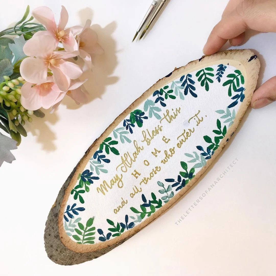 Decorative wooden plaque with a handwritten quote surrounded by painted green and blue leaves, with pink flowers and a hand holding the plaque.