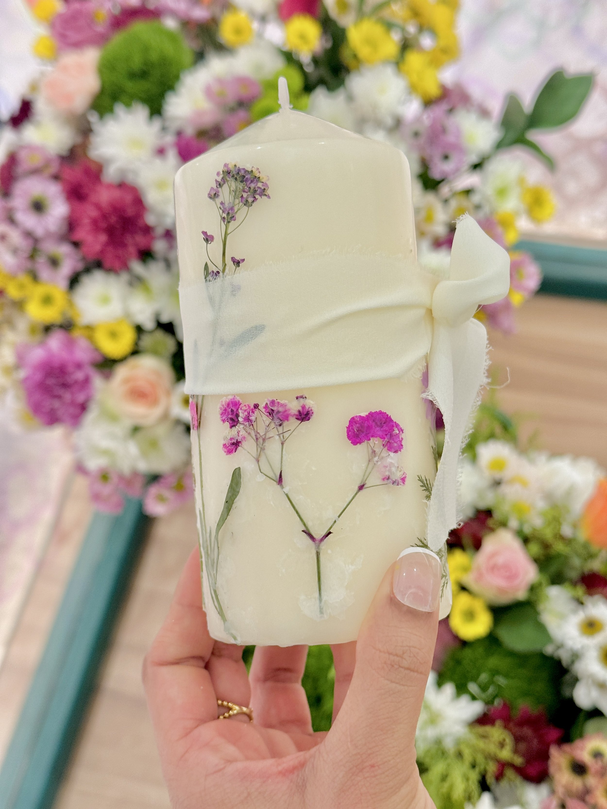 A hand holding a bar of soap with pressed dried flowers, in front of a colorful flower display.
