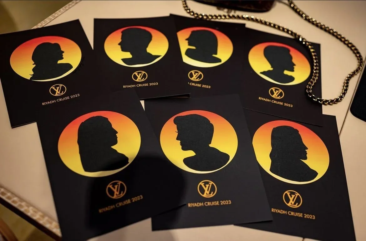 Multiple black cards with silhouettes of faces in profile and sunset backgrounds for Riyadh Cruise 2023, alongside a gold chain.