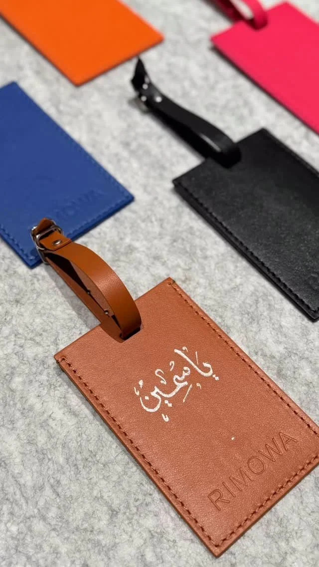 Colorful leather luggage tags in orange, pink, blue, black, and brown, with some tags featuring Arabic script and others having the brand 'RIMOWA' engraved.