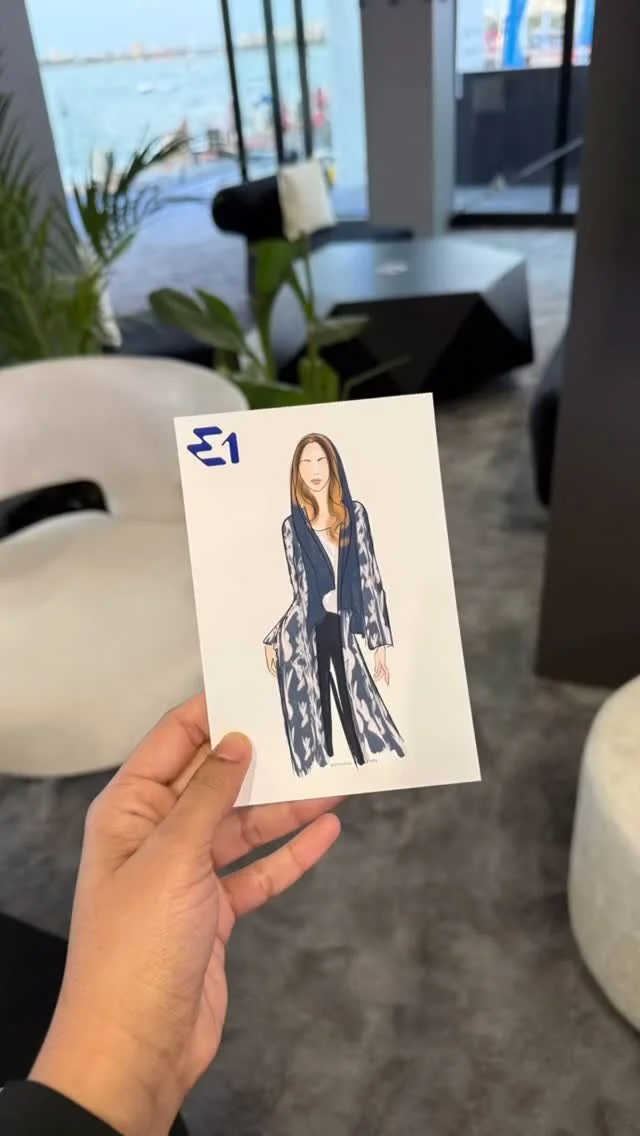 Hand holding a magazine page with a fashion illustration of a woman in a long patterned coat, standing in a modern lounge with large windows overlooking a cityscape and water.