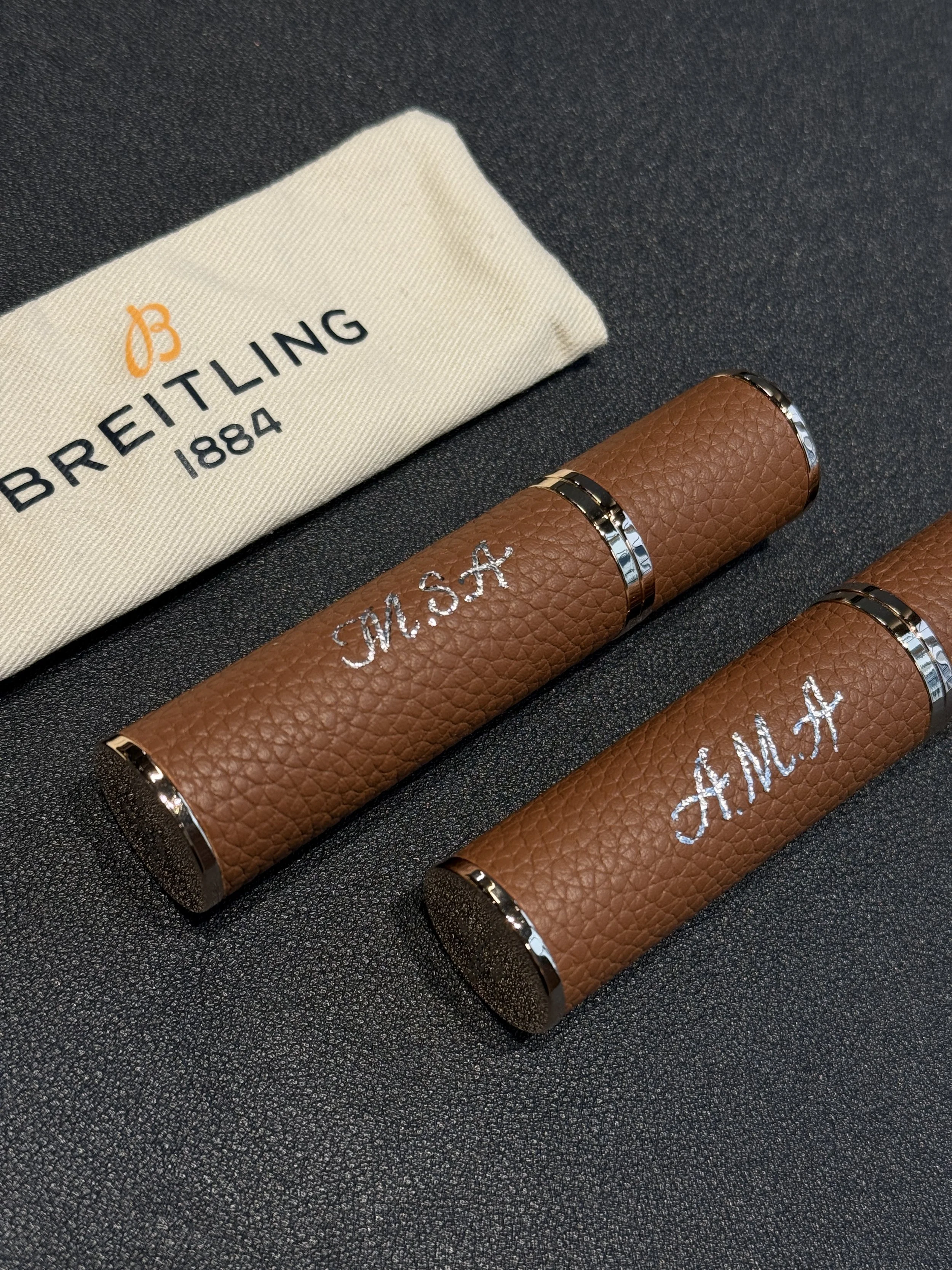 Two brown leather cylindrical objects with silver accents and embroidered initials 'M.S.A.' on a black textured surface, with a cream-colored cloth featuring the logo 'Breitling 1884' nearby.