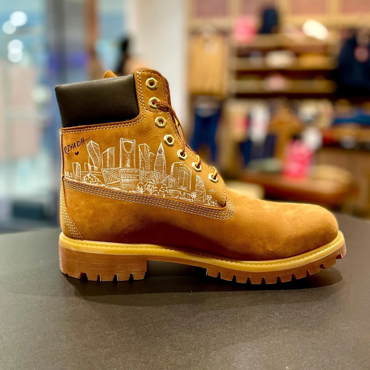 A tan work boot with a cityscape illustration and the word "RYADH" written on the side, placed on a black surface in a store environment.