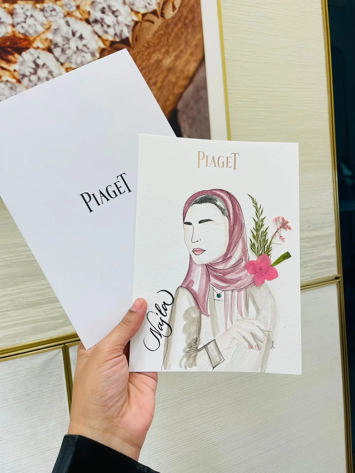 A hand holding a piece of paper with a watercolor portrait of a woman wearing a pink headscarf, with flowers and greenery, and the name "Nadja" written on it. Behind it, there are two pieces of paper with the word "PIAGET" on each. The background sho