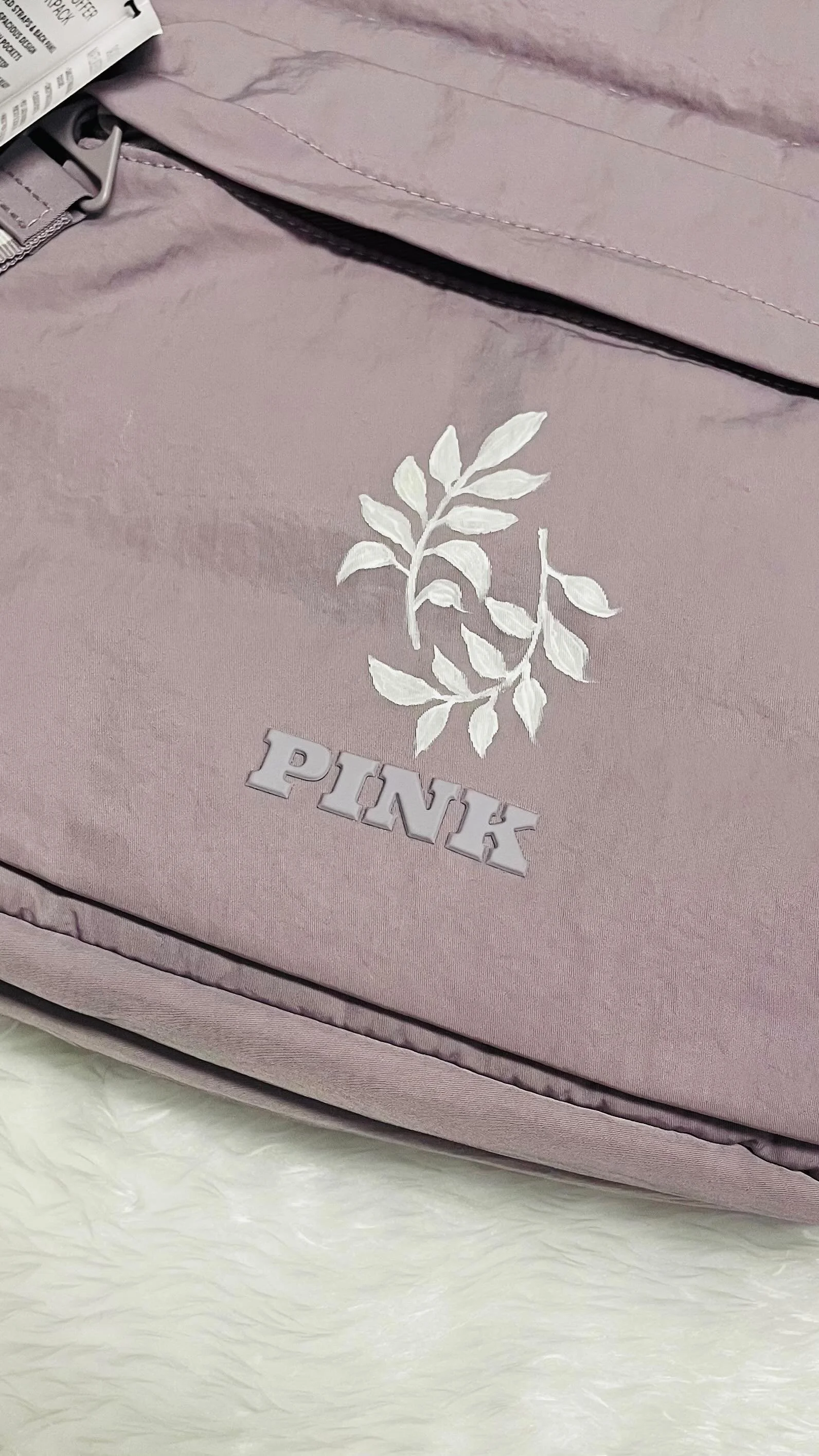 Close-up of a pale pink Victoria's Secret PINK duffel bag with white embroidered leaves and the word 'PINK' in raised letters. The bag has a zipper and a partial tag visible. The surface below is a fluffy white material.