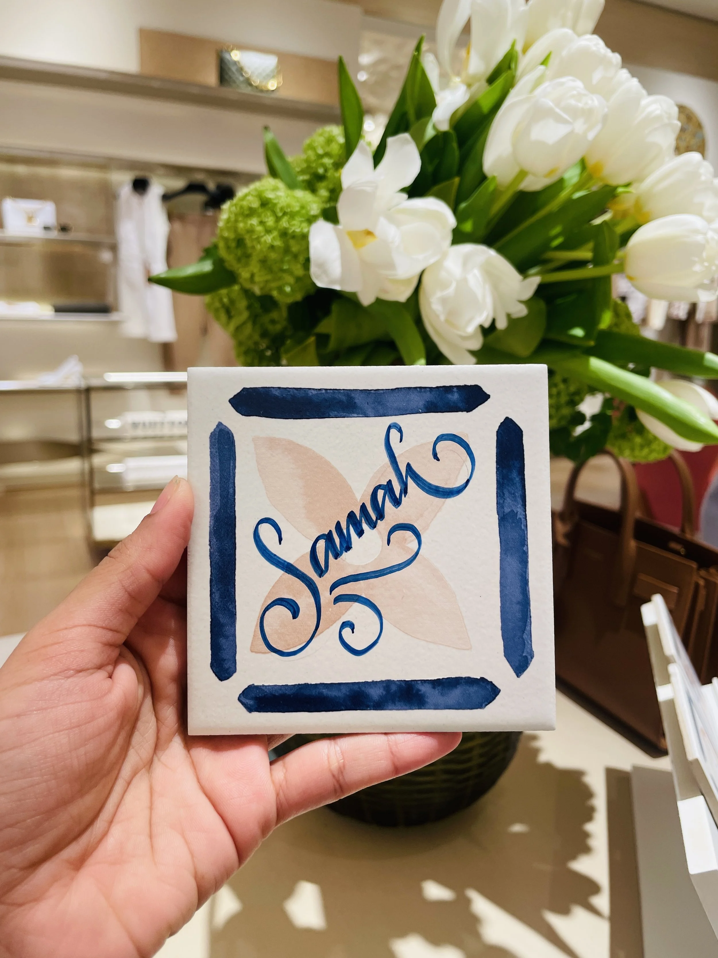 Close-up of a hand holding a white card with blue decorative border and handwritten text 'Saturday 25' in a store display area with flowers in the background.