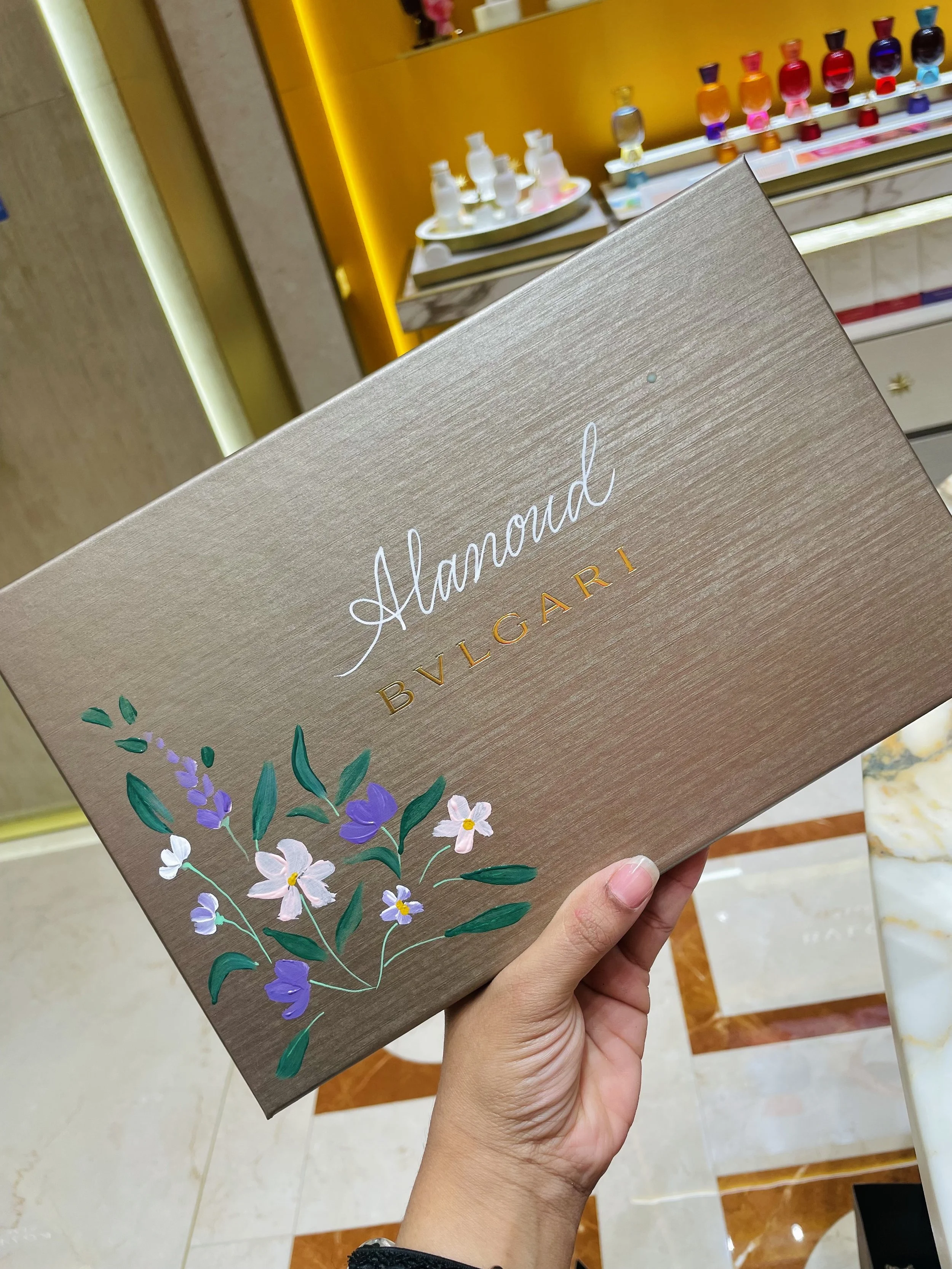 Hand holding a brochure labeled 'Alamond Bulgari' with floral illustrations on the cover, in a store with shelves of bottled perfumes in the background.