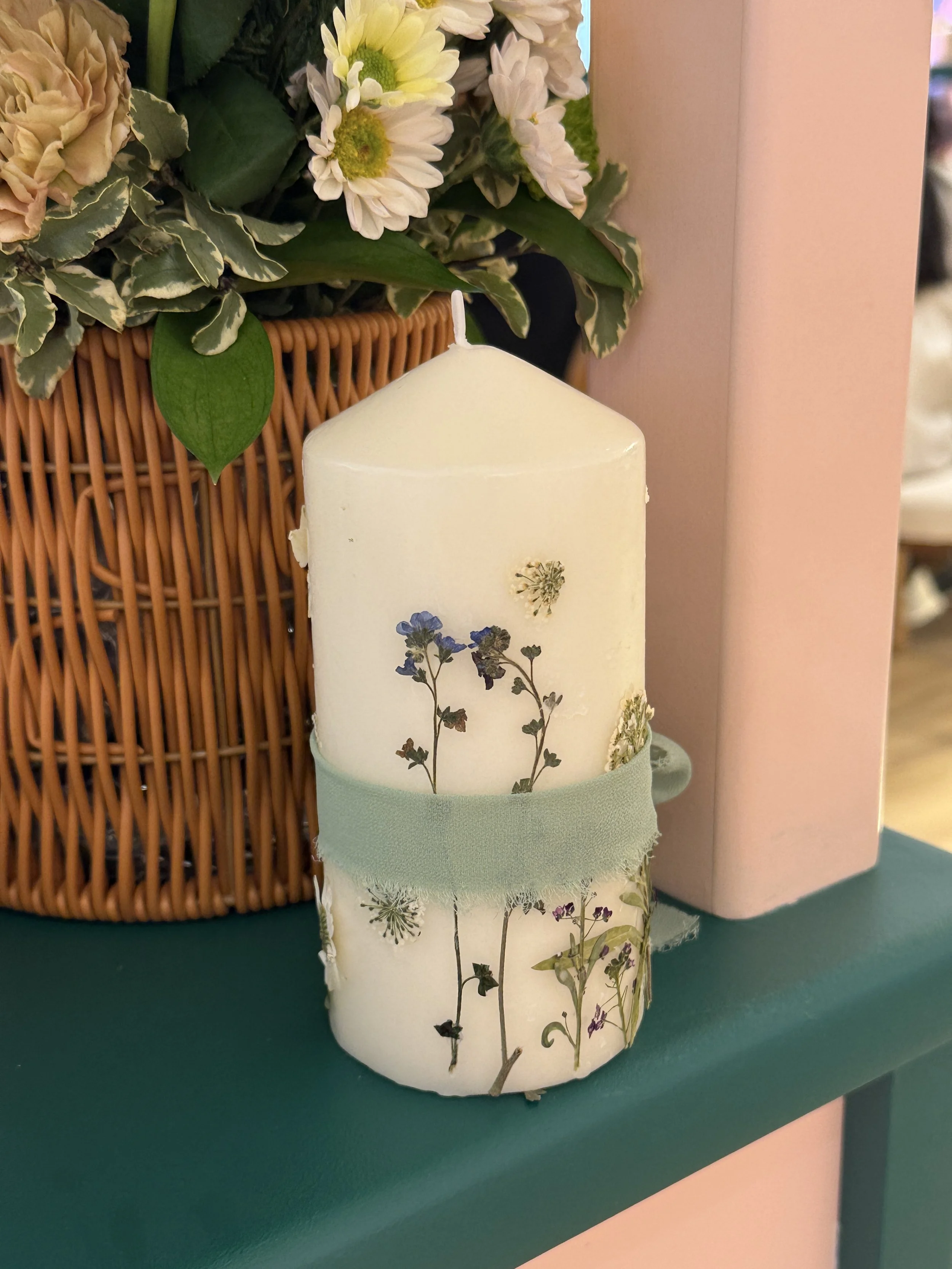 A decorated pillar wrapped in floral-patterned paper, placed on a green surface, in front of a basket of flowers.