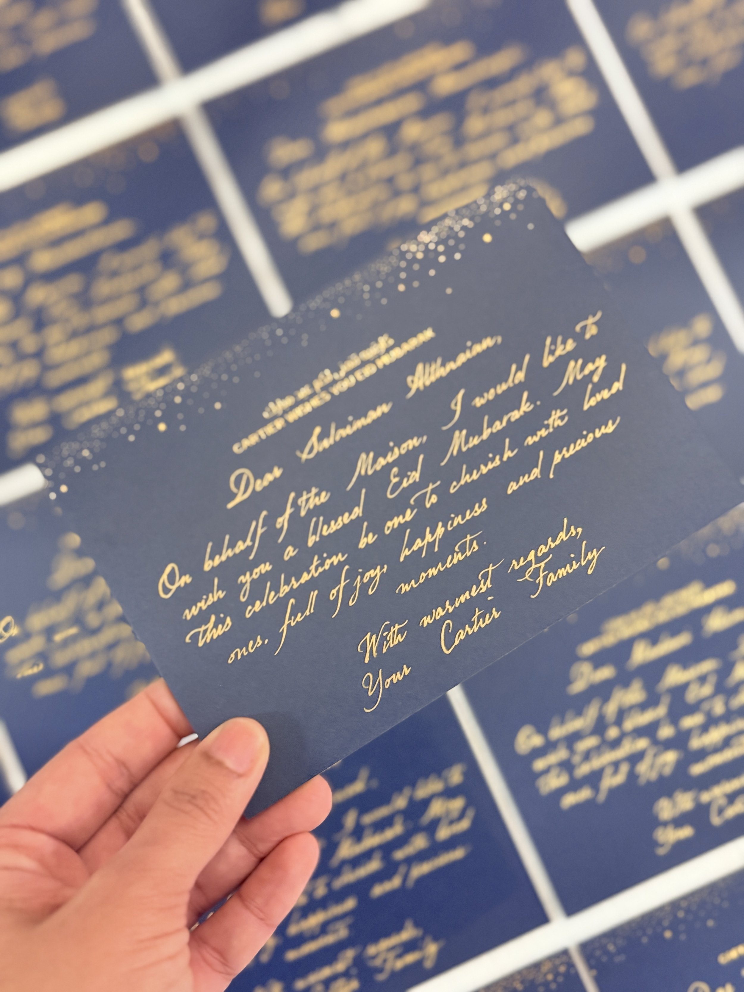 Close-up of a handwritten note in gold ink on navy blue paper, with multiple sheets stacked, held by a person. The note expresses holiday wishes and happiness.