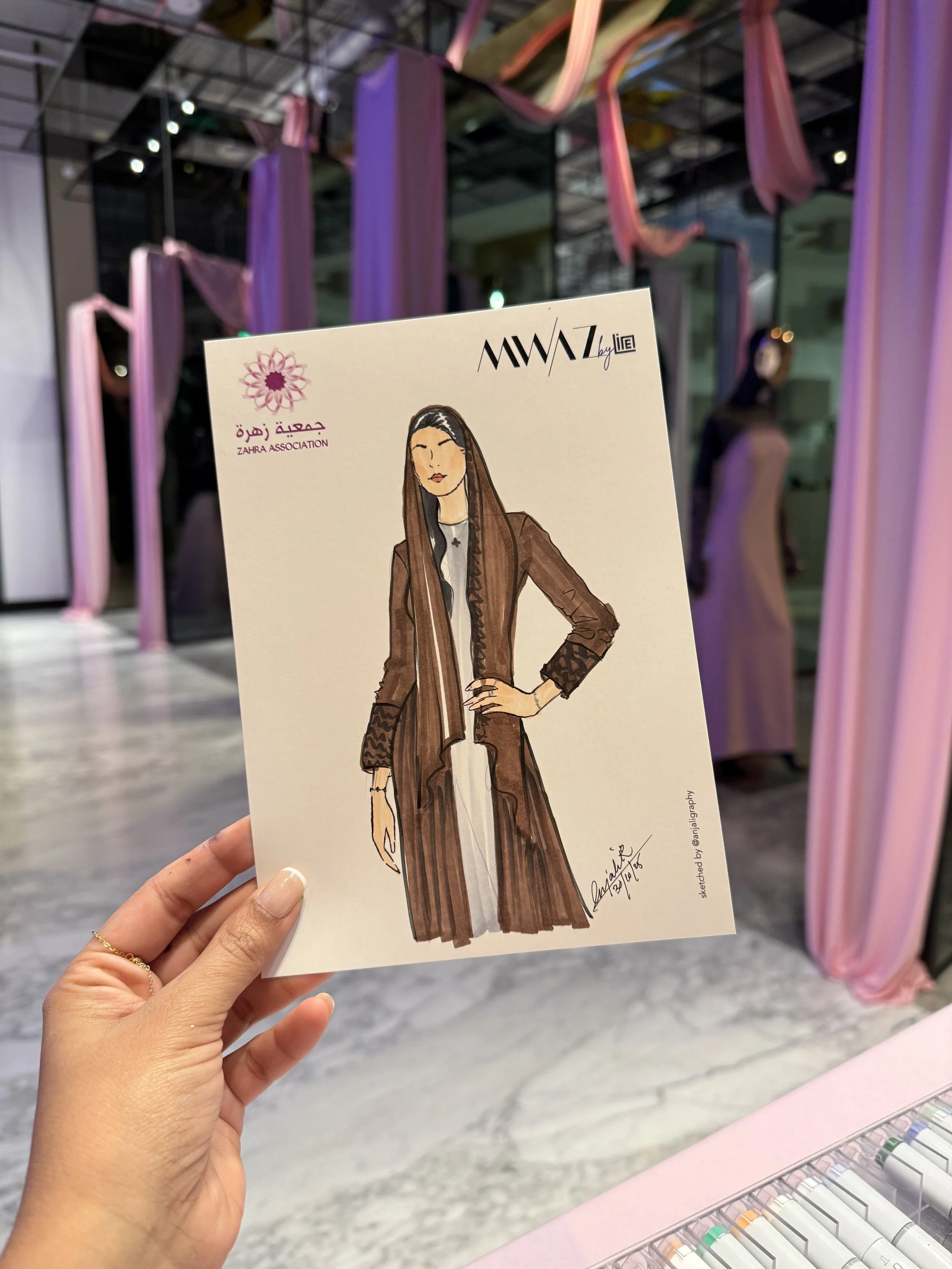 A hand holding a fashion illustration of a woman in a brown coat and long brown skirt, with pink and purple fabric art installations in the background at an art exhibit or gallery.