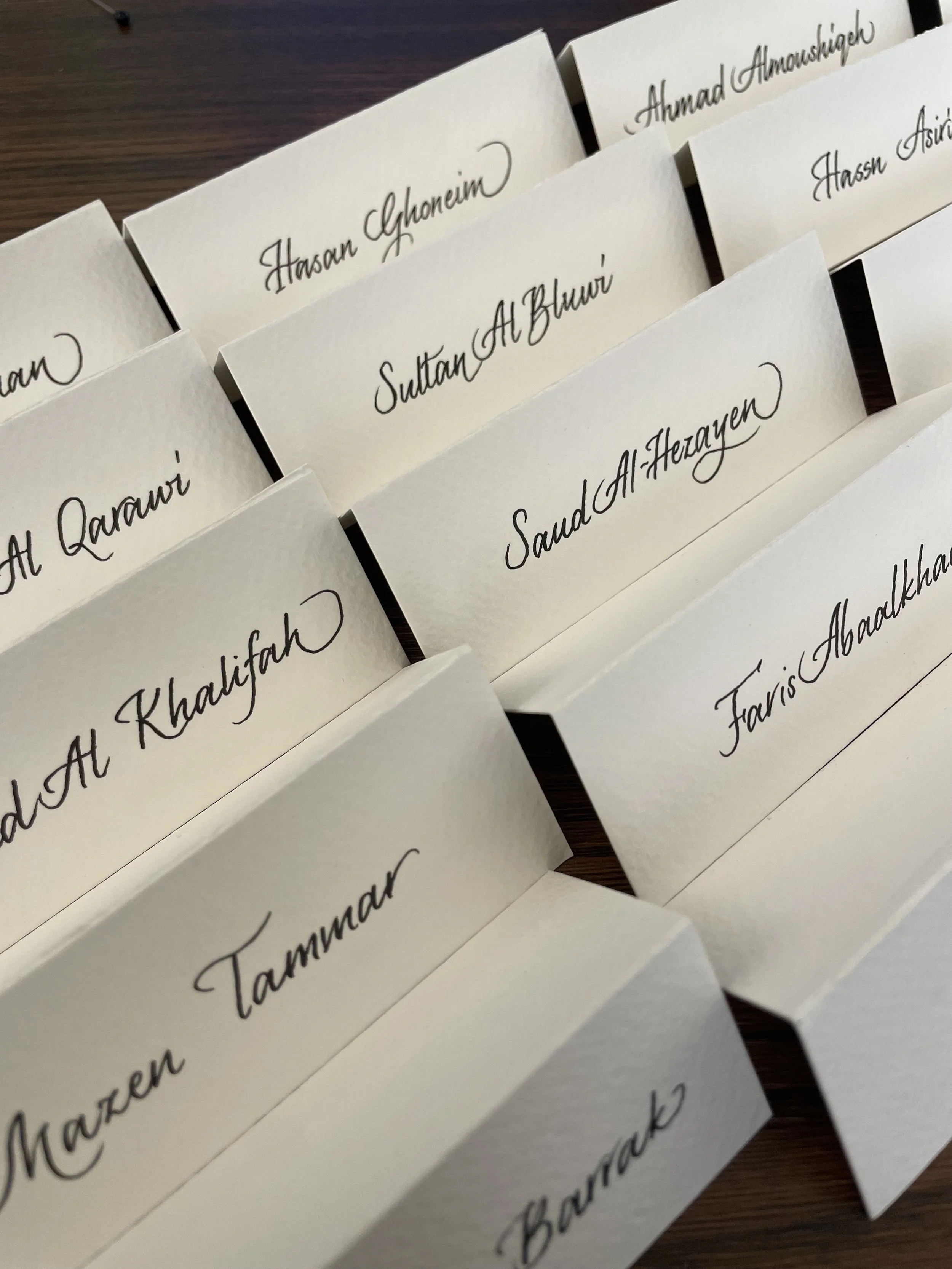 Multiple white name cards with black cursive handwriting, each with a different name, arranged on a dark wooden surface.