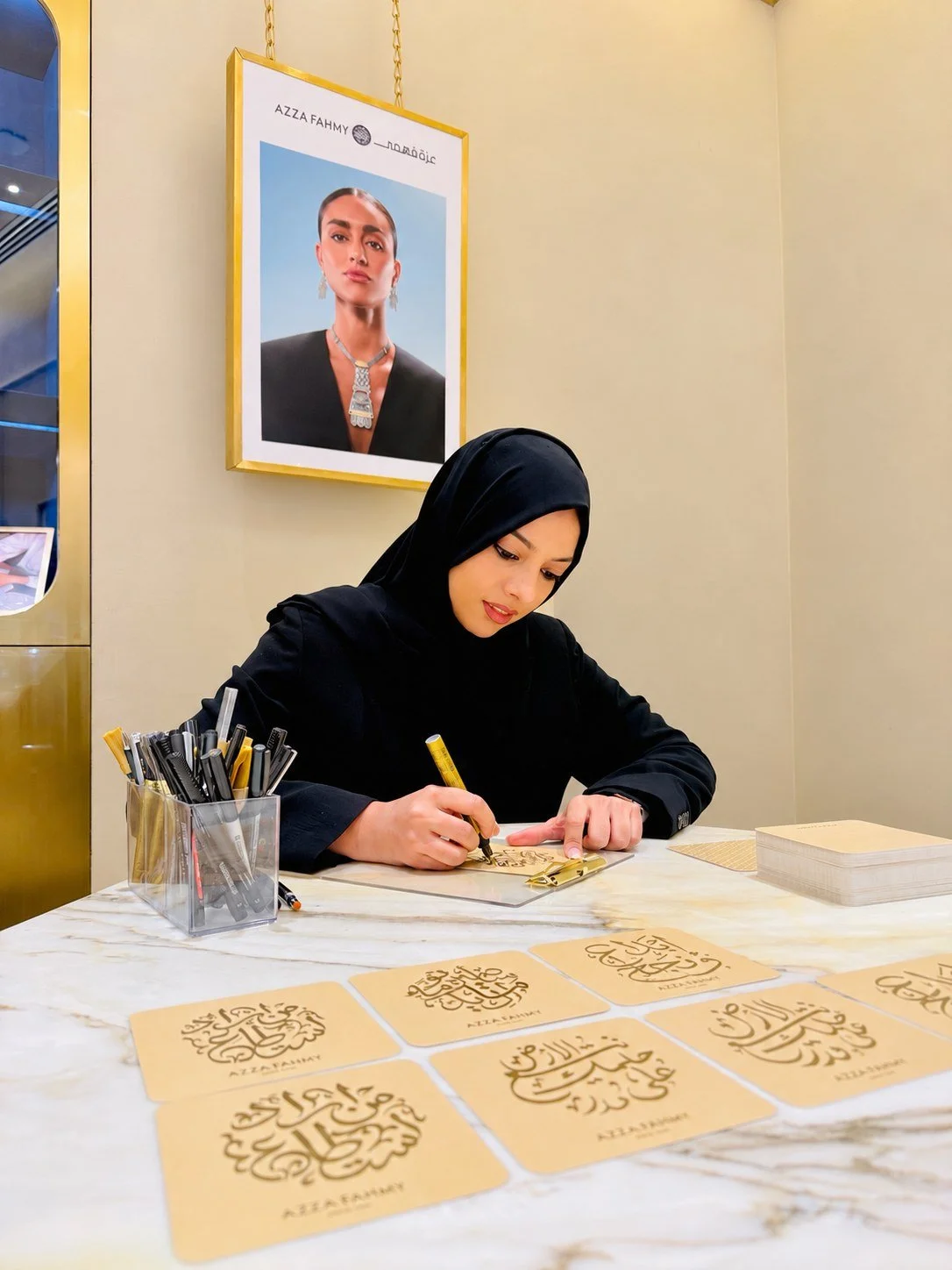 Woman dressed in black hijab and attire sitting at a marble table, creating calligraphy with black pen on gold-colored paper. Several finished calligraphy sheets are spread out on the table, and a plastic container filled with markers and pens is on 