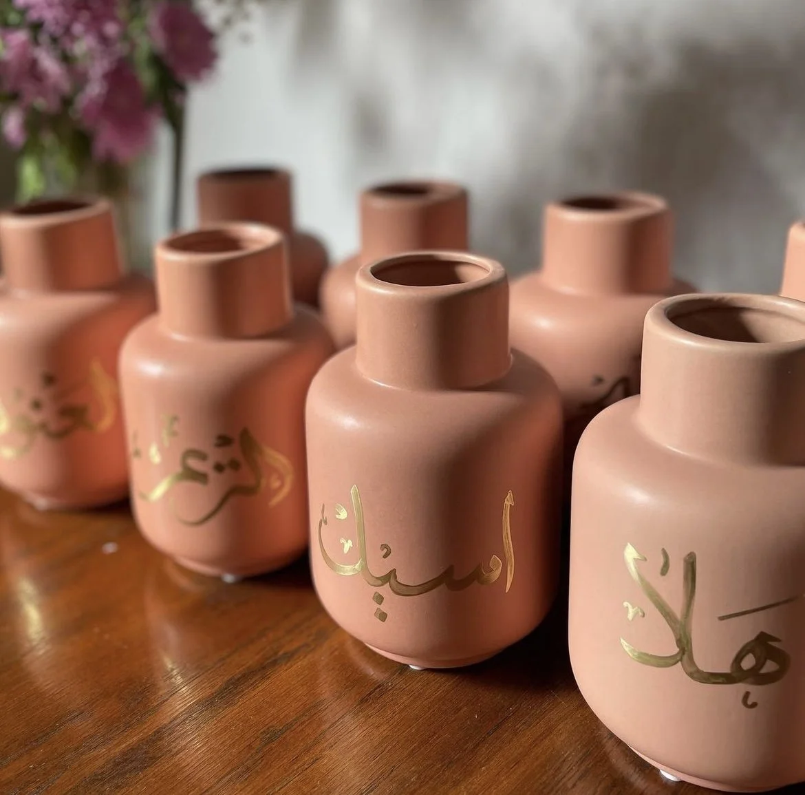 Several pink vases with gold Arabic calligraphy on a wooden surface, with a blurred background of wall and pink flowers.