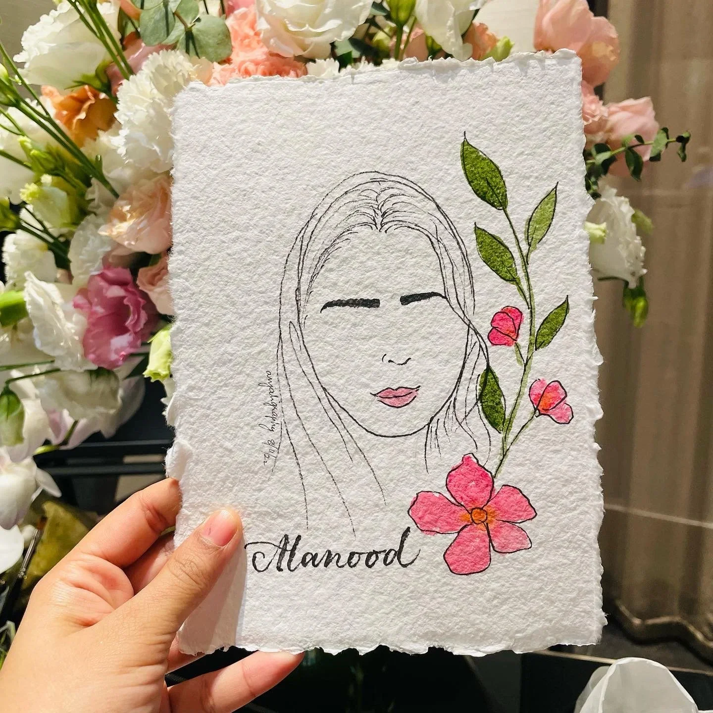 Hand-holding a piece of textured paper with a simple line drawing of a woman's face with closed eyes, pink lips, and long hair. The drawing is decorated with colorful flowers and green leaves on the right side. The word "Manood" is written in cursive