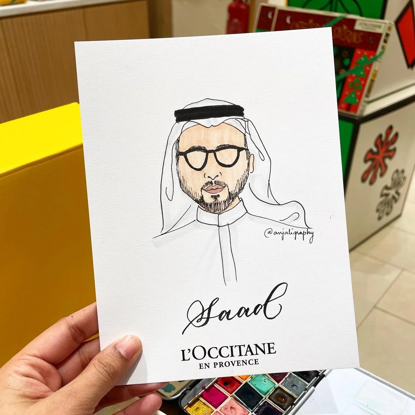 Line drawing of a man in traditional Middle Eastern attire, wearing glasses and a beard, on a white paper with the text "Saad" and "L'OCCITANE EN PROVENCE."