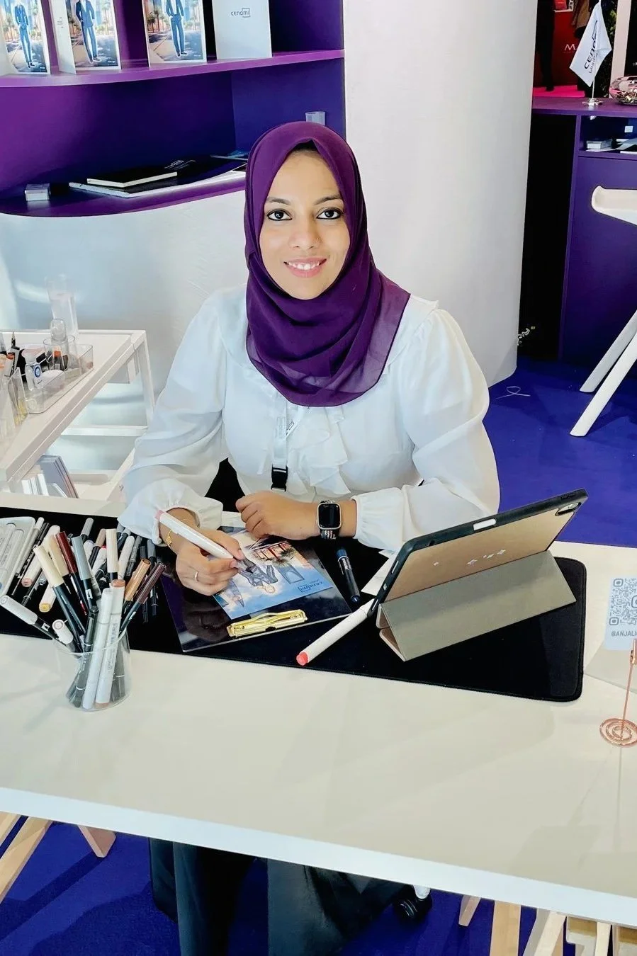 A woman wearing a purple hijab and white blouse smiling at the camera while sitting at a desk with markers, documents, and a tablet in a colorful office space.