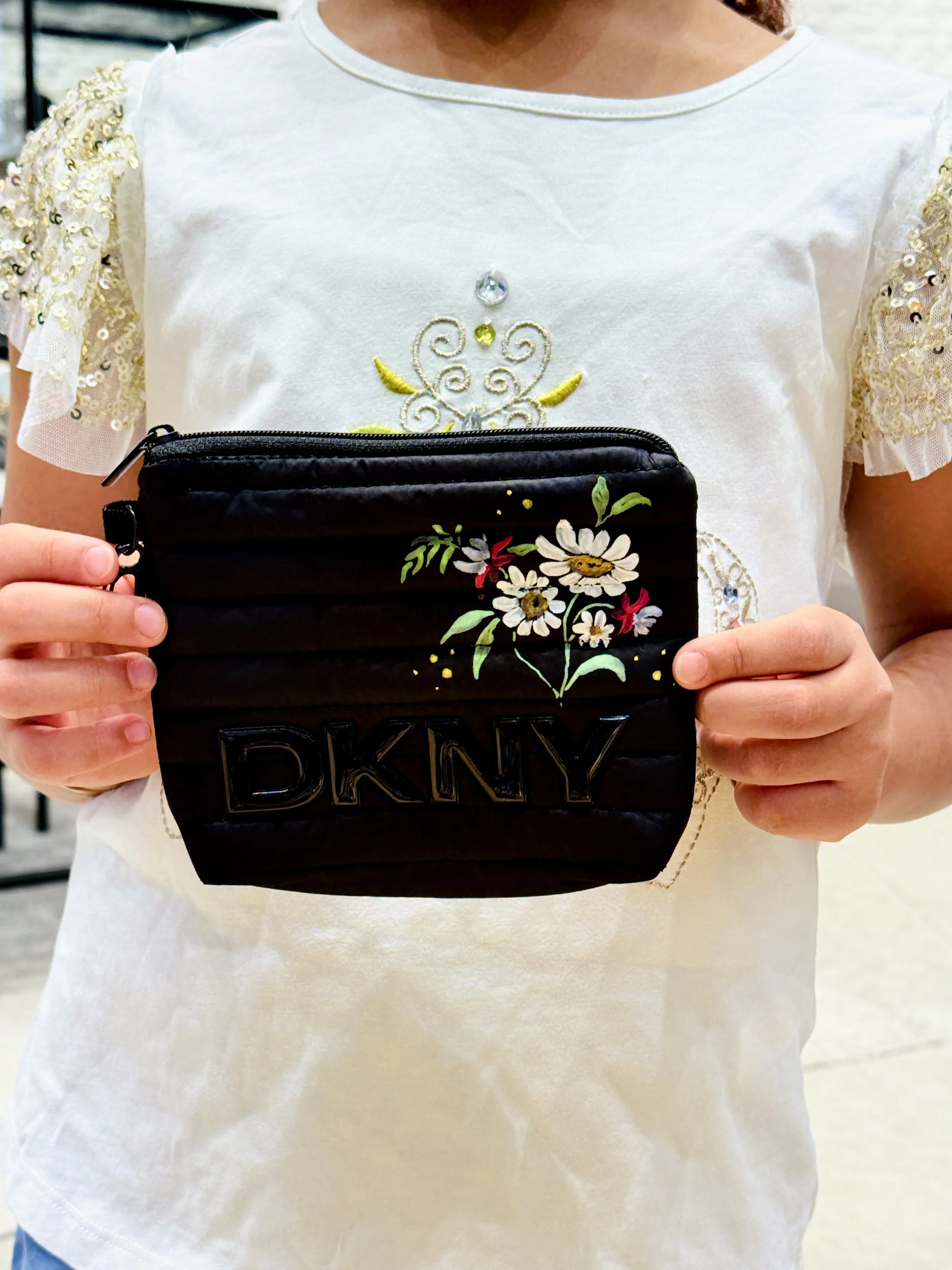 Person holding a black quilted DKNY clutch purse with floral embroidery in front of their white embroidered shirt. The shirt has decorative details on the sleeves and front.