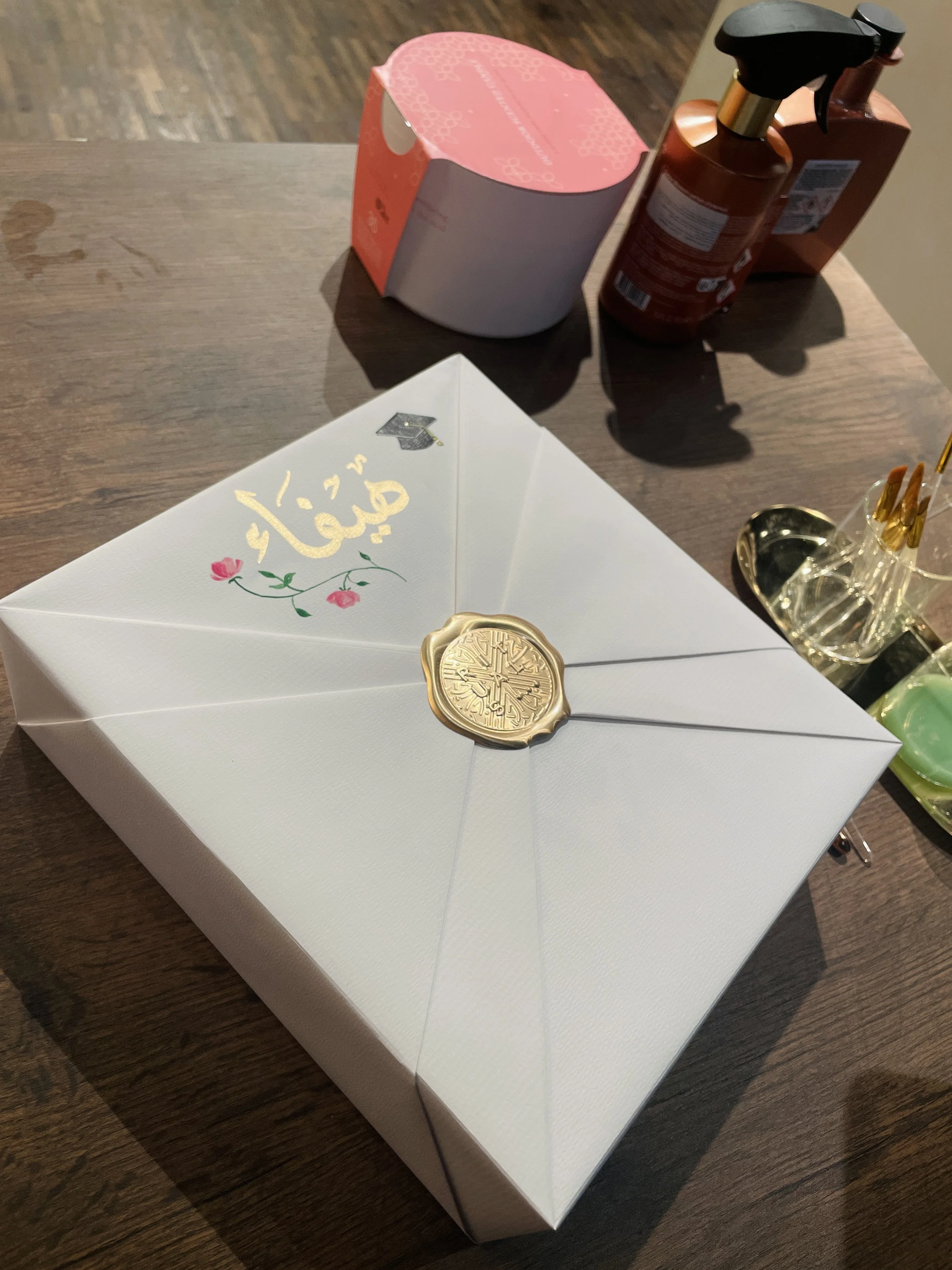 A white envelope with gold lettering and an illustration of a graduation cap and pink flowers, sealed with a gold wax seal, on a wooden surface. In the background, pink and brown bottles and a clear container with pens are visible.