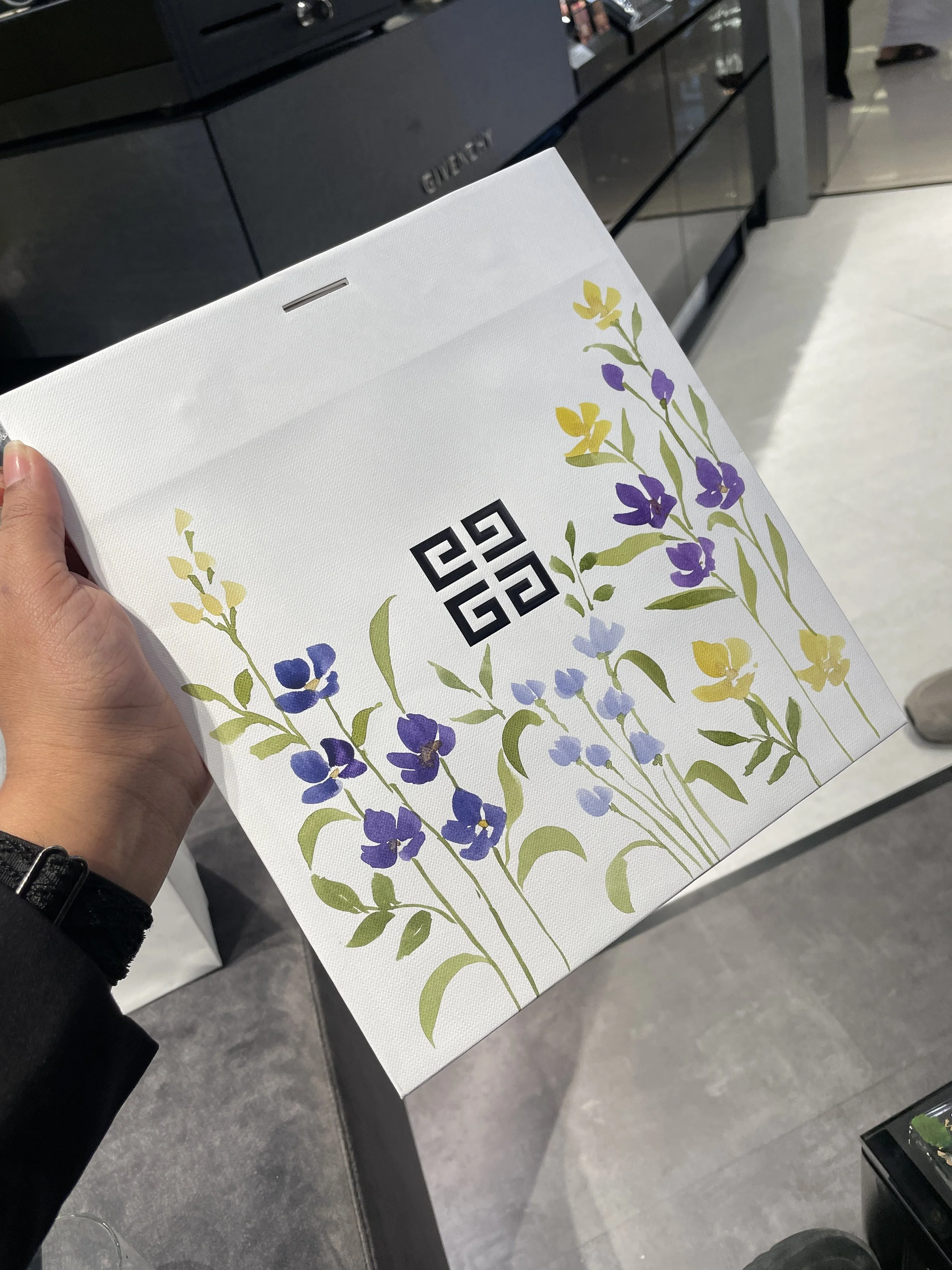 A person holding a white shopping bag with floral designs and a black logo in a retail store, with black cabinets and a person in the background.