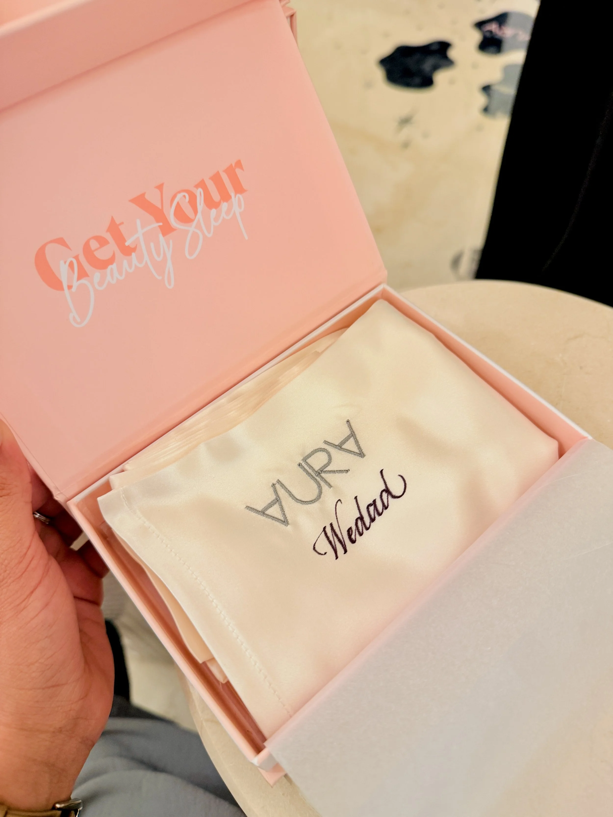 The image shows a pink box with the words "Get Your Beauty Sleep" printed on the inside of the lid. Inside the box is a beige cloth with embroidered text that reads "LUXURY Bedding." The background includes a beige table and some laundry on the floor