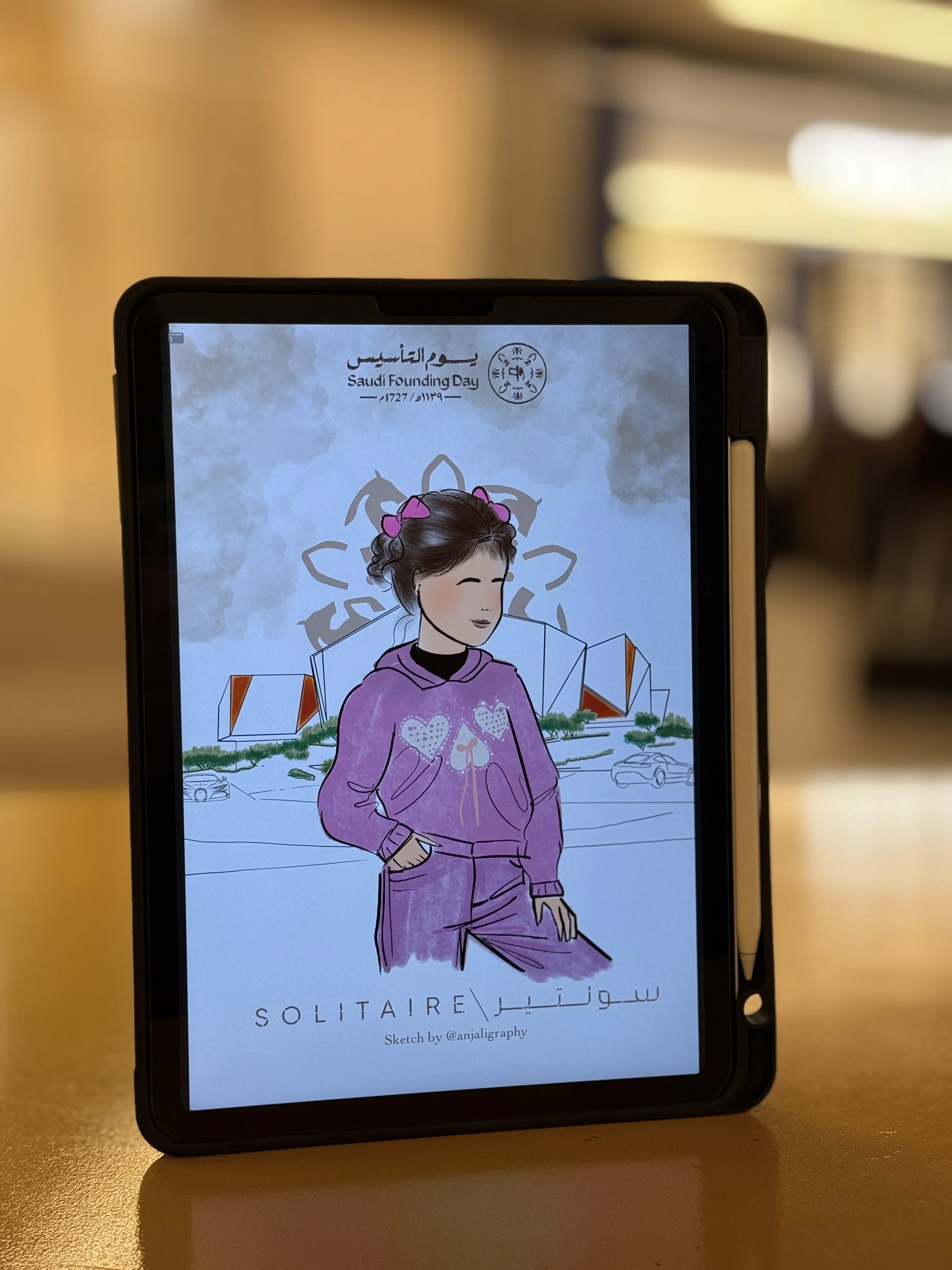 A digital screen displays a drawing of a woman with dark hair tied with pink bows, wearing a purple hoodie with heart designs, set against a cityscape. The text above reads 'Saudi Founding Day' in both Arabic and English with the years 1727 and 2023.