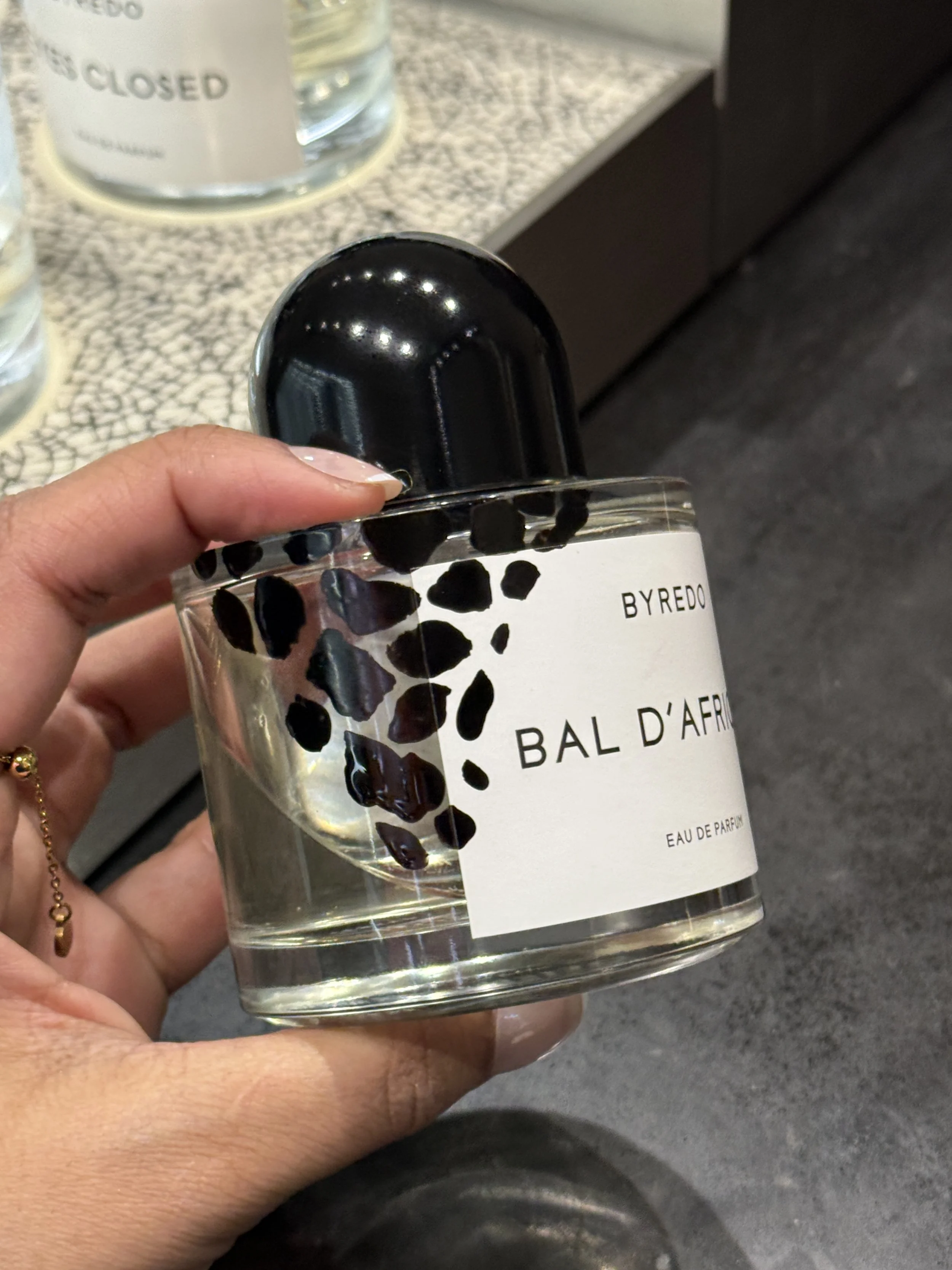 A person holding a clear glass perfume bottle with black stones embedded in the liquid. The bottle has a black cap and a white label that reads 'BYREDO BAL D'AFR' and 'EAU DE PARFUM'. In the background, there is a counter with glass jars, one marked 