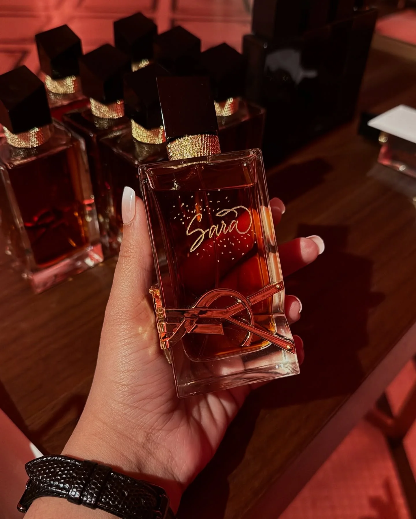 A hand holding a personalized glass perfume bottle with the name 'Sara' written on it, in front of a display of similar perfume bottles on a wooden surface.